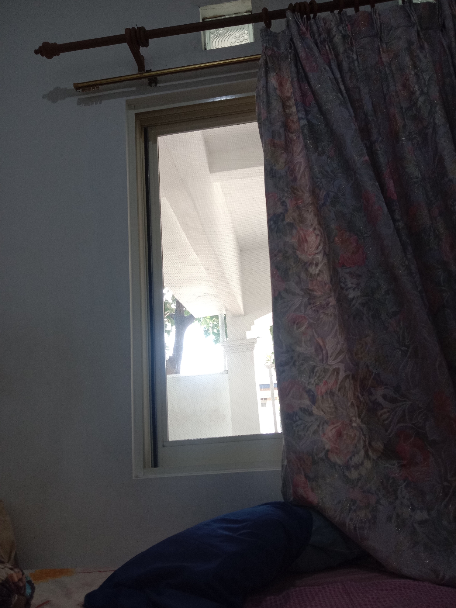 The image depicts a room with a window on the right side, partially covered by a patterned curtain. The window frame is light beige. Outside the window, a light-colored structure, possibly a porch or balcony, is visible, with a tree and more architectural elements in the background. A dark blue pillow rests on a surface covered with a pink and white patterned cloth in the foreground. A double curtain rod is mounted above the window, with a reddish-brown rod on top and a thinner gold rod below. The walls of the room are painted a light gray. The scene appears to be indoors, likely a bedroom, with the lighting suggesting it is daytime. The location is identified as Ngingas, Indonesia.