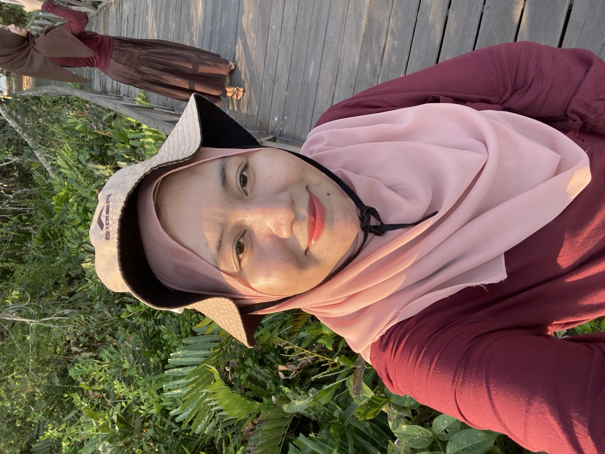 The image shows a selfie taken by a woman in the foreground, who is wearing a pink hijab, a maroon long-sleeved shirt, and a light-colored bucket hat. She is smiling at the camera. Behind her, on a wooden boardwalk, another woman wearing a brown hijab and a long brown skirt is standing and looking away from the camera while holding a phone. The environment appears to be a natural setting with dense green foliage to the left of the boardwalk and the wooden planks of the boardwalk stretching into the distance. The lighting suggests it is either sunrise or sunset. The location is identified as Sungai Pitung, Indonesia.