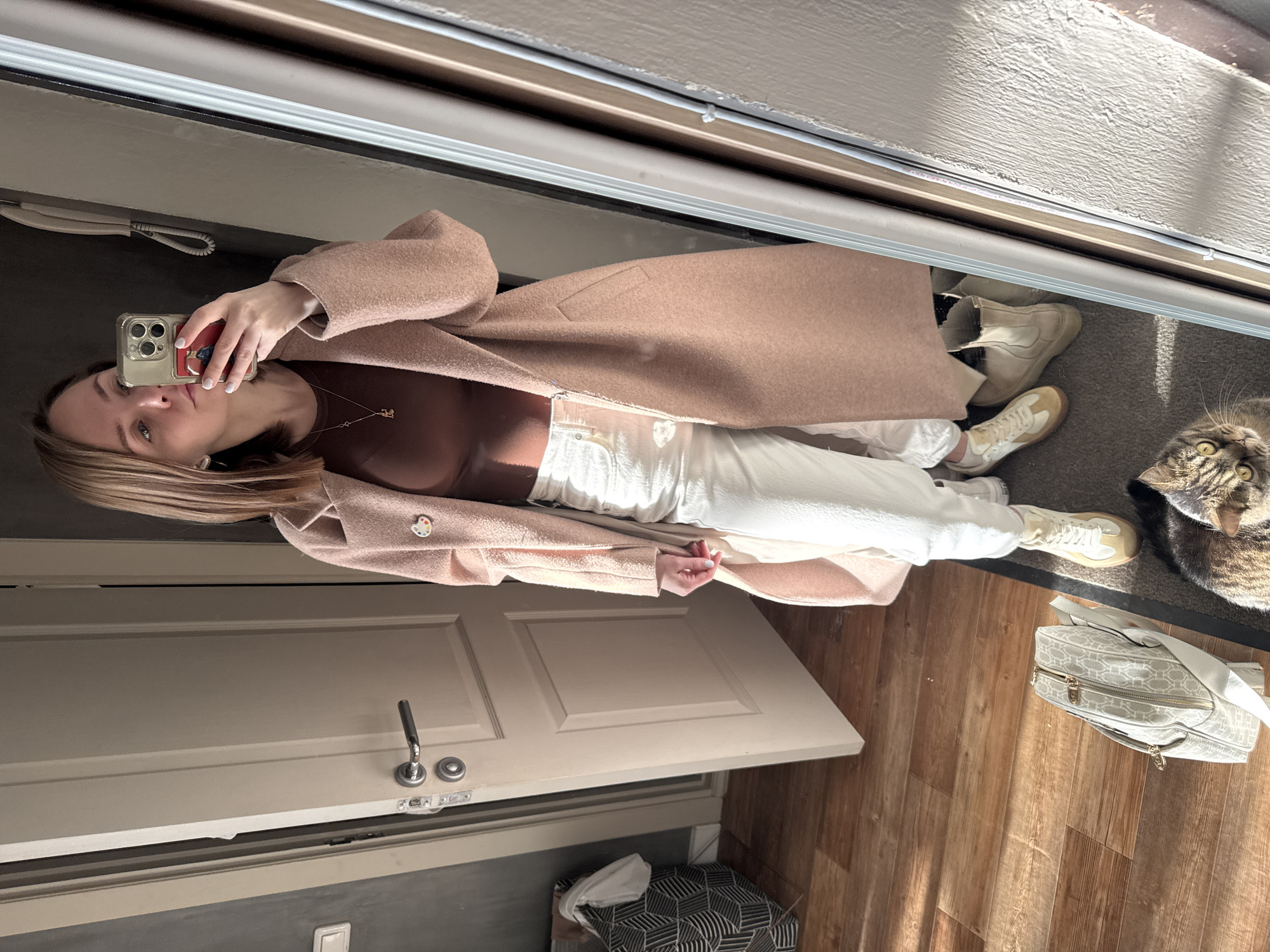 A woman takes a selfie in a full-length mirror. She is wearing a brown top, white pants, a light brown coat, and sneakers. A cat is partially visible on the floor to the right of the woman, along with a patterned bag. The location appears to be an interior entryway with a closed door and wooden flooring. The city of Perm, Russia is not evident in this image.