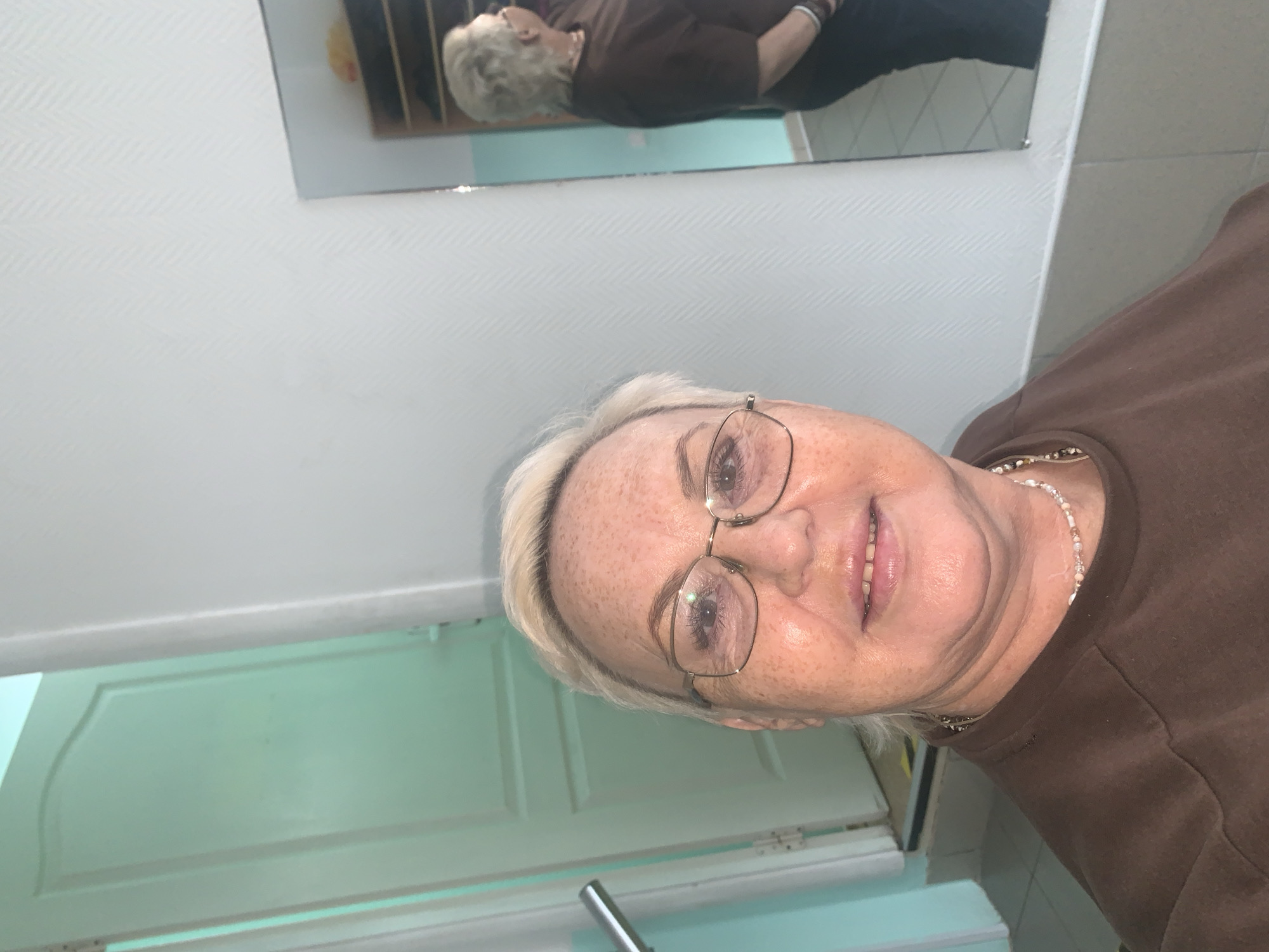 A selfie shows a woman with short, blonde hair and glasses. She is wearing a brown shirt and a beaded necklace. A mirror in the background reflects an image of the same woman, wearing a similar outfit. She seems to be standing in a bathroom with a white door. The location appears to be indoors, possibly in a residence, but there is no information to suggest the city of Chaikovsky, Russia.