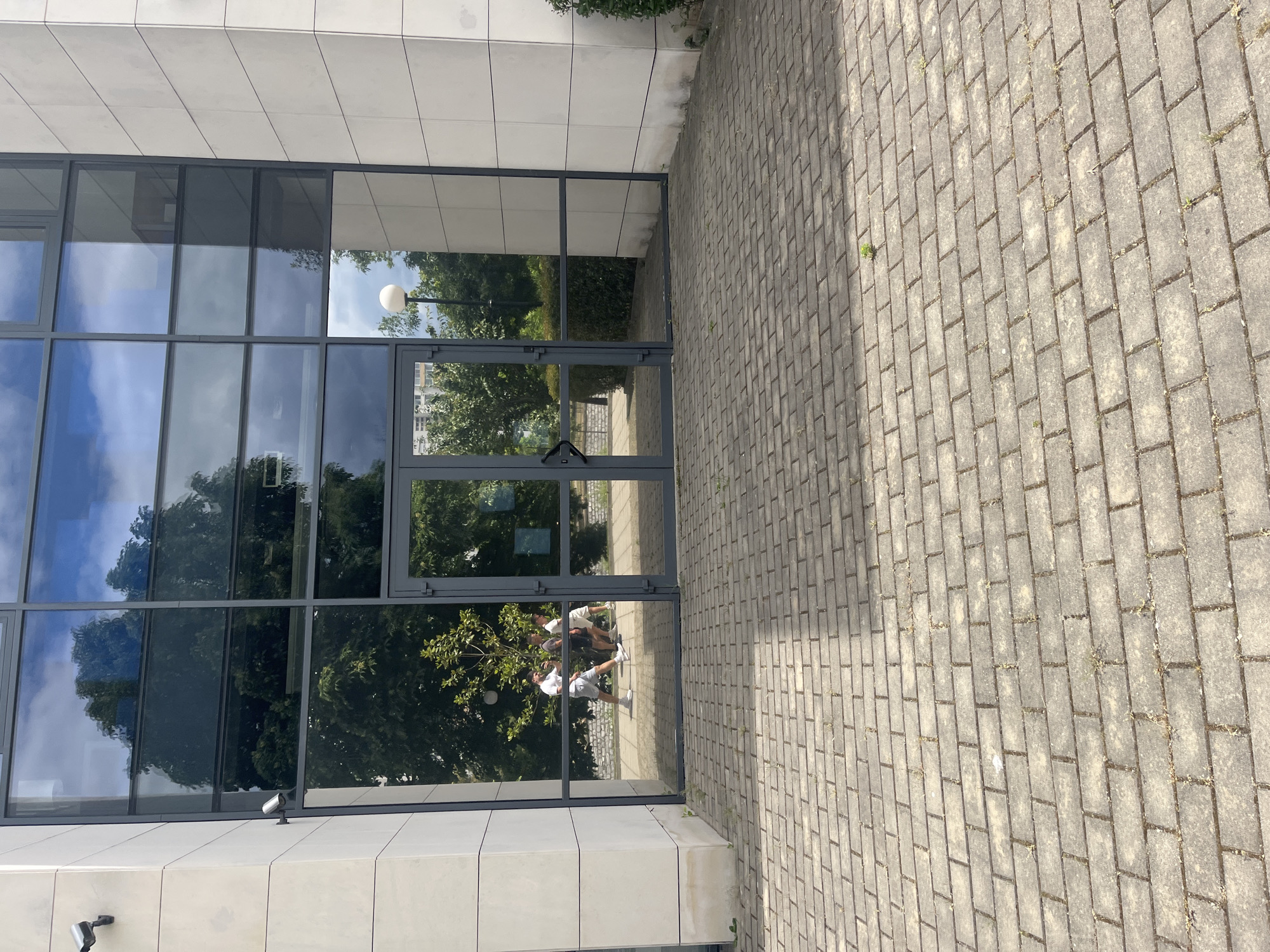 A modern building with a large glass facade stands next to a paved sidewalk. The glass reflects the sky, trees, and a lamppost. The reflection also shows two people. The location is suspected to be in Blagoevgrad, Bulgaria.