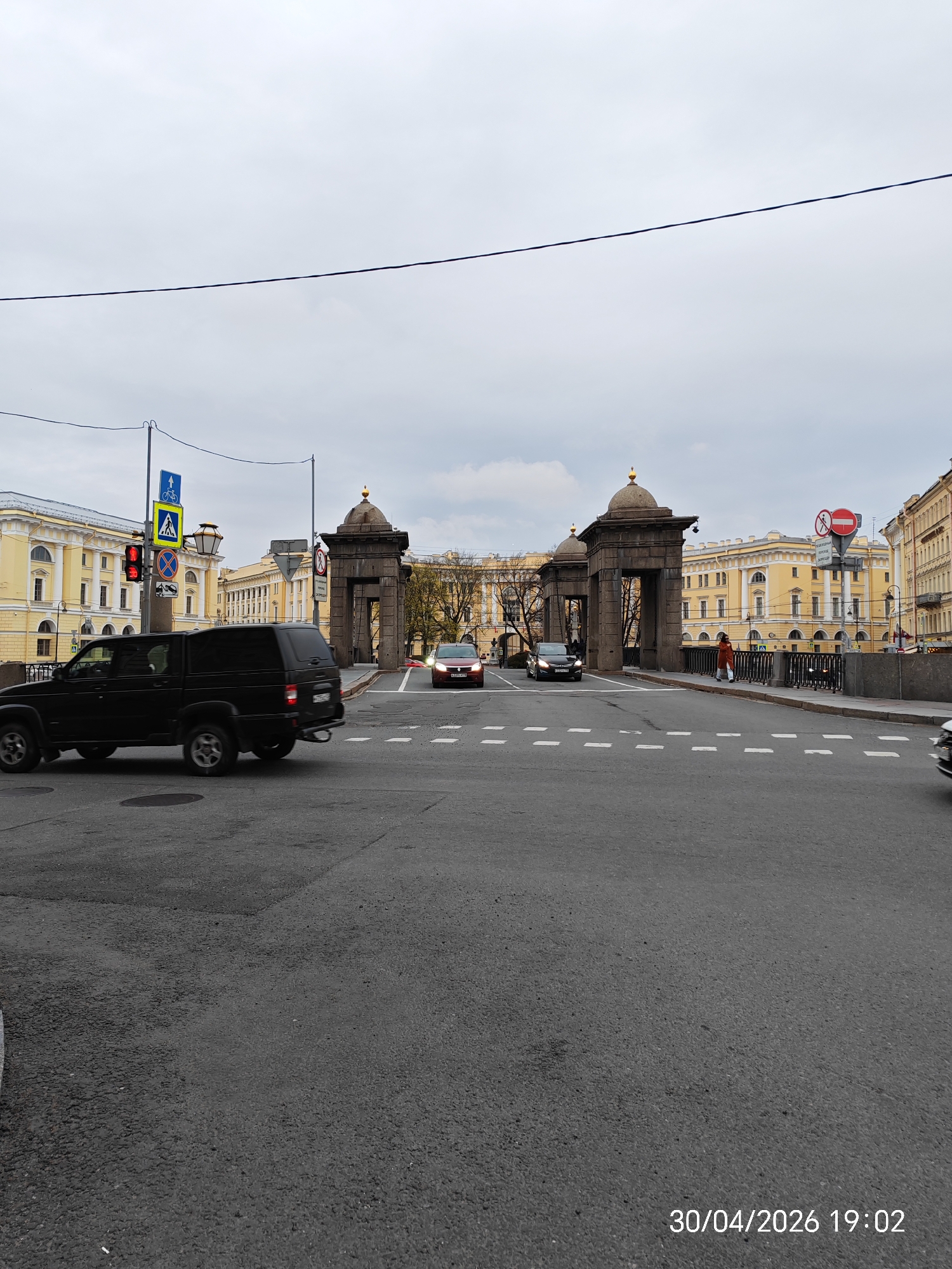 The image depicts an urban street scene centered around a bridge with distinctive architectural elements, identifiable as the Lomonosov Bridge in St. Petersburg, Russia. Two large, grey stone gate towers with golden-domed cupolas stand prominently on the bridge, framing the view down the street.
In the foreground, a black SUV is positioned on the left side of the road, appearing to turn left or proceed from an intersection. Its design suggests a rugged vehicle. Further back on the bridge, a dark sedan is visible, and a red car is seen approaching from the distance. A white vehicle is partially visible on the far right. A single pedestrian, dressed in a red coat, is walking on the sidewalk of the bridge near the right gate tower.
The surrounding buildings are large, classical in style, painted a distinct yellow, with multiple stories and arched windows. The sky is overcast, suggesting a cloudy day, though the timestamp "30/04/2026 19:02" indicates it is early evening.
Visible details include several traffic signs: a traffic light showing red, a blue square sign with symbols for a pedestrian crossing and a bicycle lane, a blue square sign with a white upward arrow indicating a one-way street or straight-ahead direction, and a red circular "no entry" sign. Power lines are visible stretching across the upper portion of the frame. The asphalt road features clear white dashed lane markings.