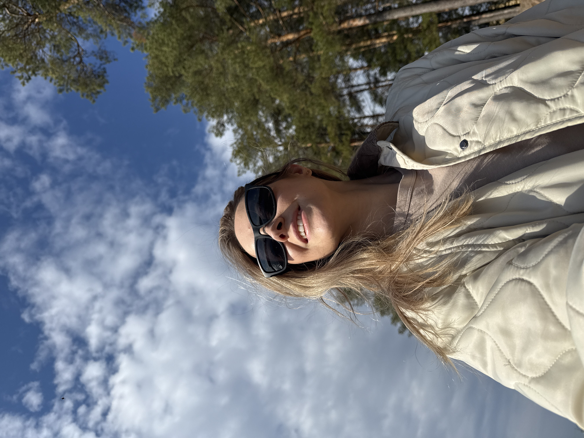 The image displays a portrait of a young woman, shown in a rotated orientation, against a backdrop of a clear blue sky and green trees. The woman is smiling, wearing black sunglasses, and has long, light-colored hair. She is dressed in a light-colored, possibly cream or off-white, quilted jacket over a lighter top. The photograph appears to be taken outdoors on a sunny day, with a clear blue sky and scattered white clouds visible above and behind her. The tops of pine trees suggest a forest or wooded setting. Her smile conveys a cheerful and relaxed mood. There is no visible text in the image. The scene is set in an outdoor environment, likely a forest or park in Chaikovsky, Russia.