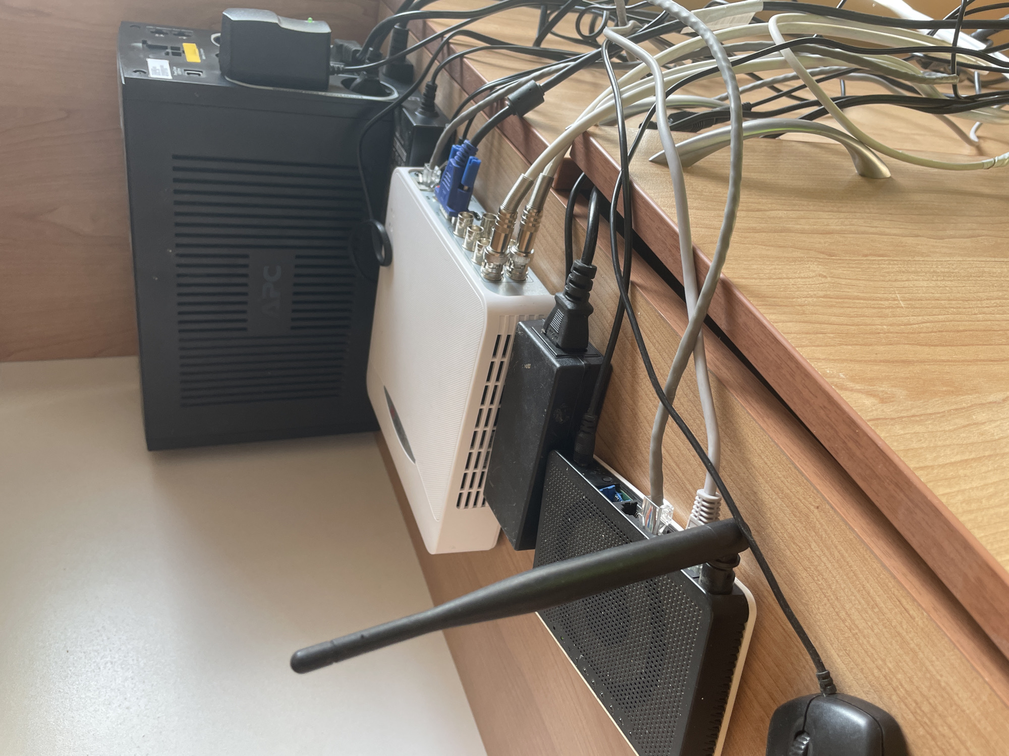The image presents a detailed view of a compact electronic equipment setup, likely in an indoor setting such as a home office or technical corner. On the left, a black Uninterruptible Power Supply (UPS) unit, branded "APC," stands vertically. A power adapter is plugged into the top of this unit.
To the right of the UPS, a white, rectangular device, possibly a Digital Video Recorder (DVR) or Network Video Recorder (NVR), is prominently featured. Its rear panel is connected with multiple coaxial cables (BNC connectors) and a blue VGA cable, suggesting video input and output capabilities, likely for surveillance. A black power brick is plugged into the right side of this white device.
Further to the right and slightly below, a black network router or modem is visible, distinguished by a long, dark antenna extending towards the viewer. A small black computer mouse is partially visible in the bottom right corner, connected by a cable.
The scene is dominated by a dense tangle of black, white, and gray cables that snake between the devices and extend upwards and out of frame, indicating numerous power and data connections. The equipment rests on a light-colored, possibly white or beige, surface, with a wooden shelf or desk forming the background. No specific activity is taking place, but the arrangement of devices suggests their continuous operation for power backup, video recording/surveillance, and network connectivity. The lighting is uniform, providing no specific time-of-day cues. The only visible text is the "APC" logo on the UPS. The location context provided is Ihtiman, Bulgaria.