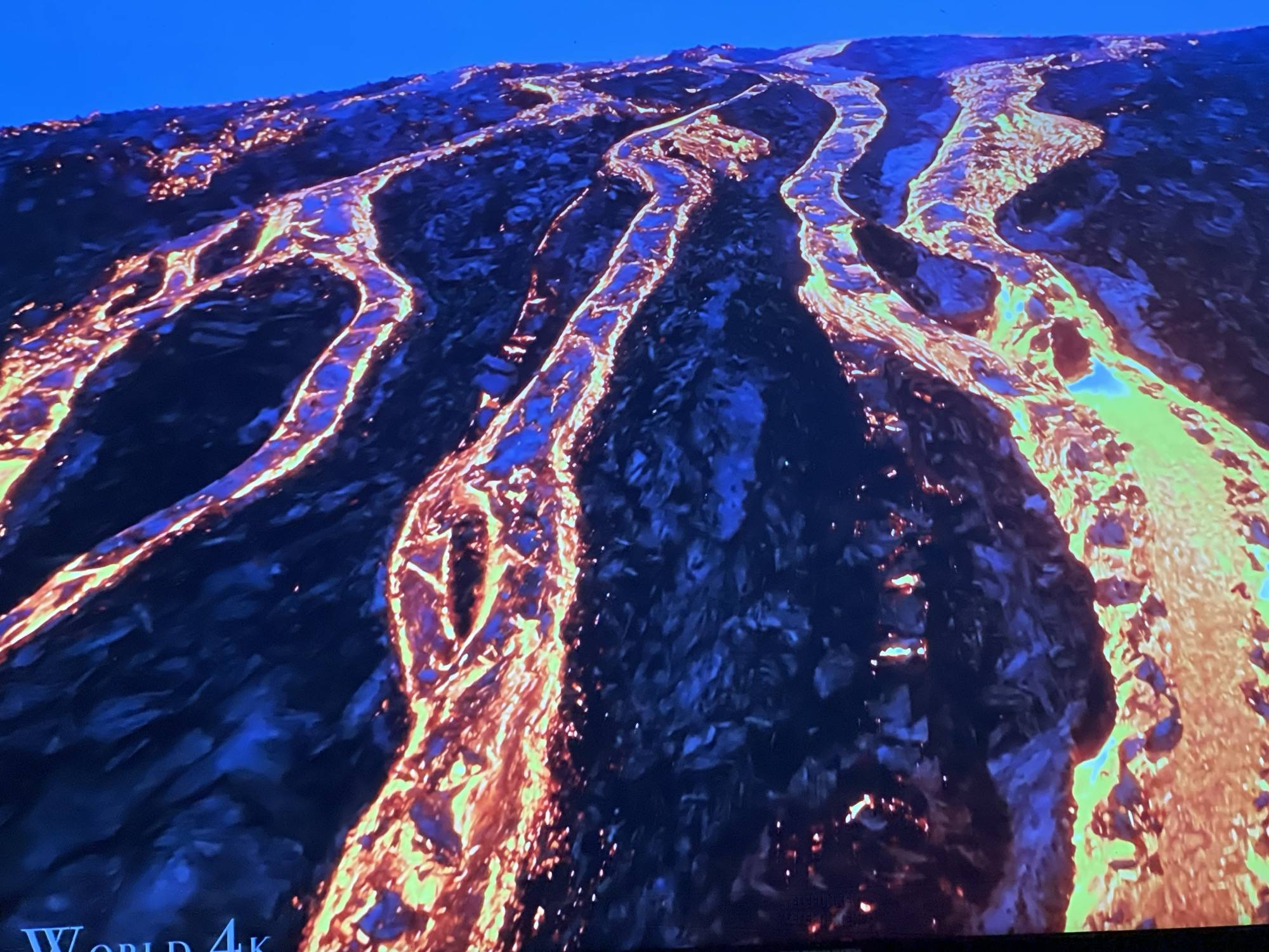 The image displays a vibrant, aerial view of flowing lava. The molten rock, rendered in fiery oranges, yellows, and greens, snakes across a dark, textured landscape. The lava appears to be in motion, with its incandescent glow illuminating the surrounding terrain. The upper portion of the image shows a clear, deep blue sky. There are no people or structures visible in the frame, and the scene depicts a natural geological event. The location is not identifiable as Sofia, Bulgaria, as there are no urban elements or specific landmarks present.