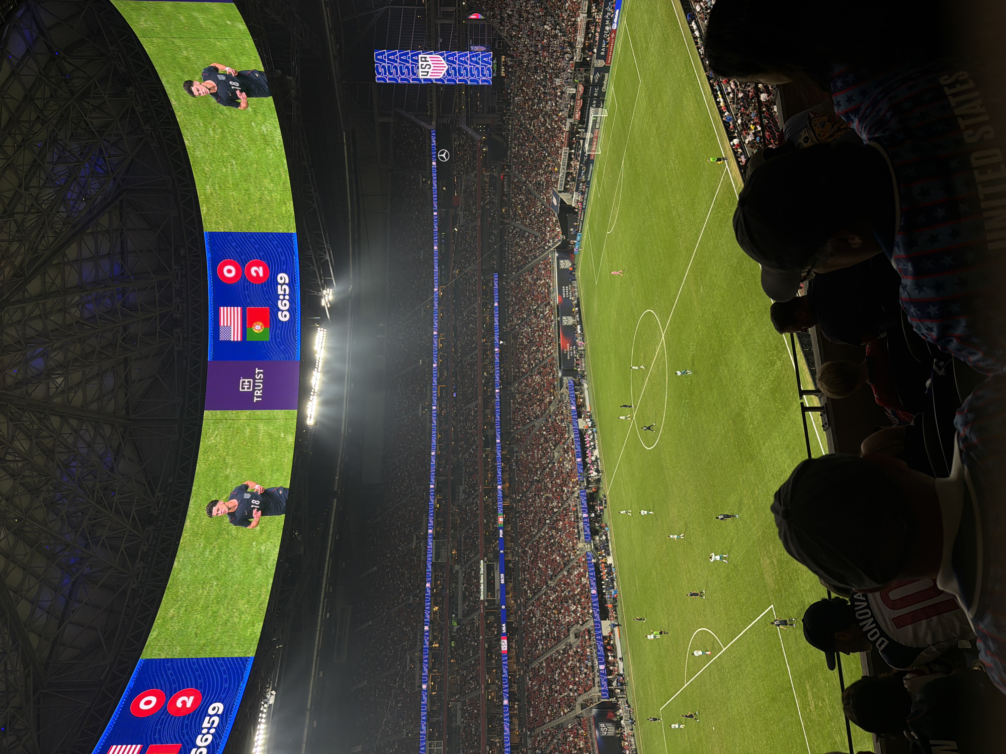 The image captures a live soccer game at Mercedes-Benz Stadium in Atlanta, United States. The stadium appears to be packed with spectators. On a large curved screen overhead, the score is displayed as USA 0 - Portugal 2. The time remaining in the game is 66:59. An image of a soccer player wearing number 18 is also shown on the screen. A young fan in the foreground is wearing a Donovan jersey with the number 10. An individual behind the fan is wearing clothing with a United States design. The soccer field is in view with the game in progress.