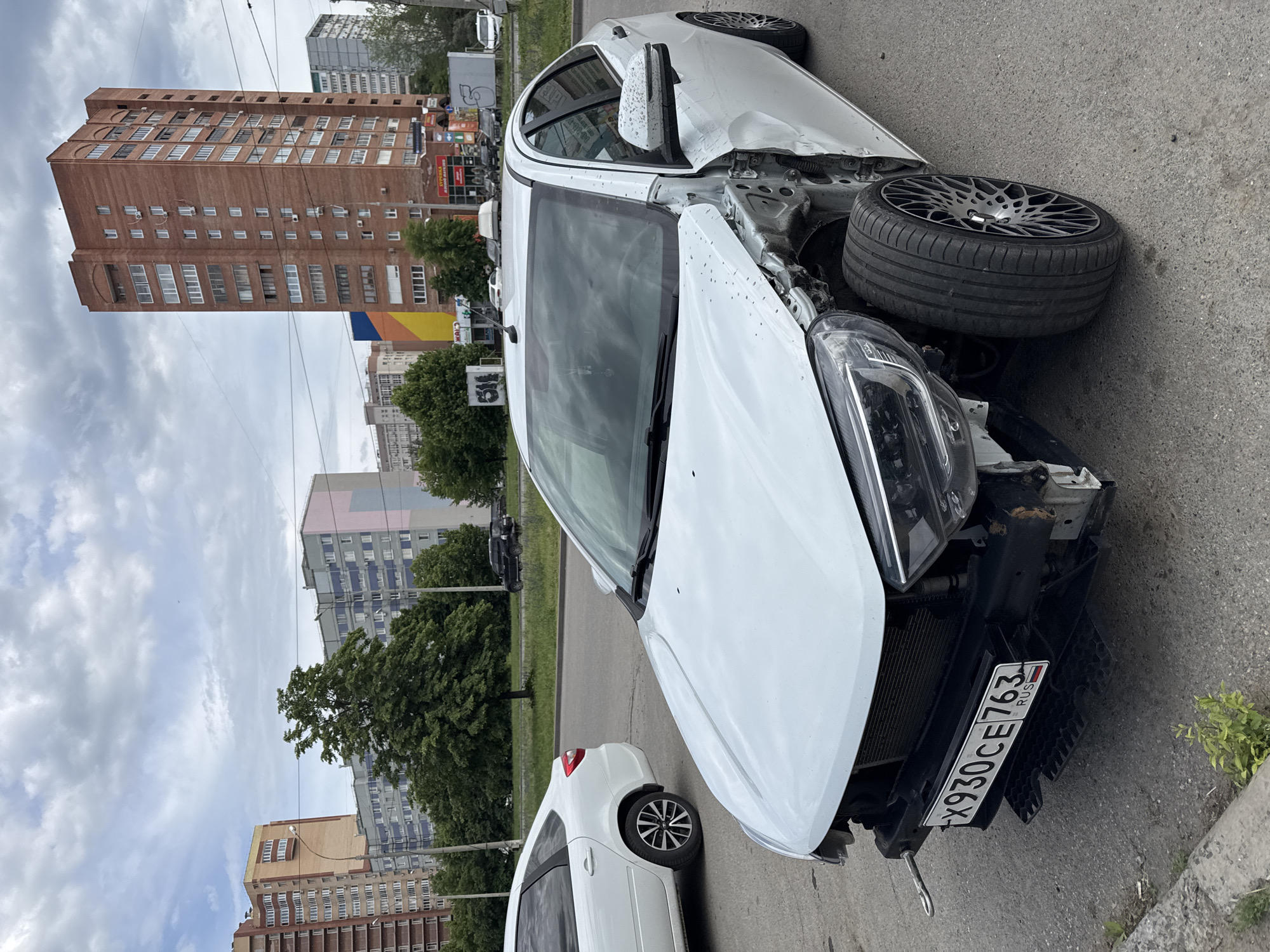 A white car with significant front-end damage is parked on a city street. The front tire is dislodged and resting on the hood. The car's license plate reads "X930CE763 RUS." Other cars are parked nearby, and apartment buildings form the urban backdrop. The location is Unknown, Unknown.
