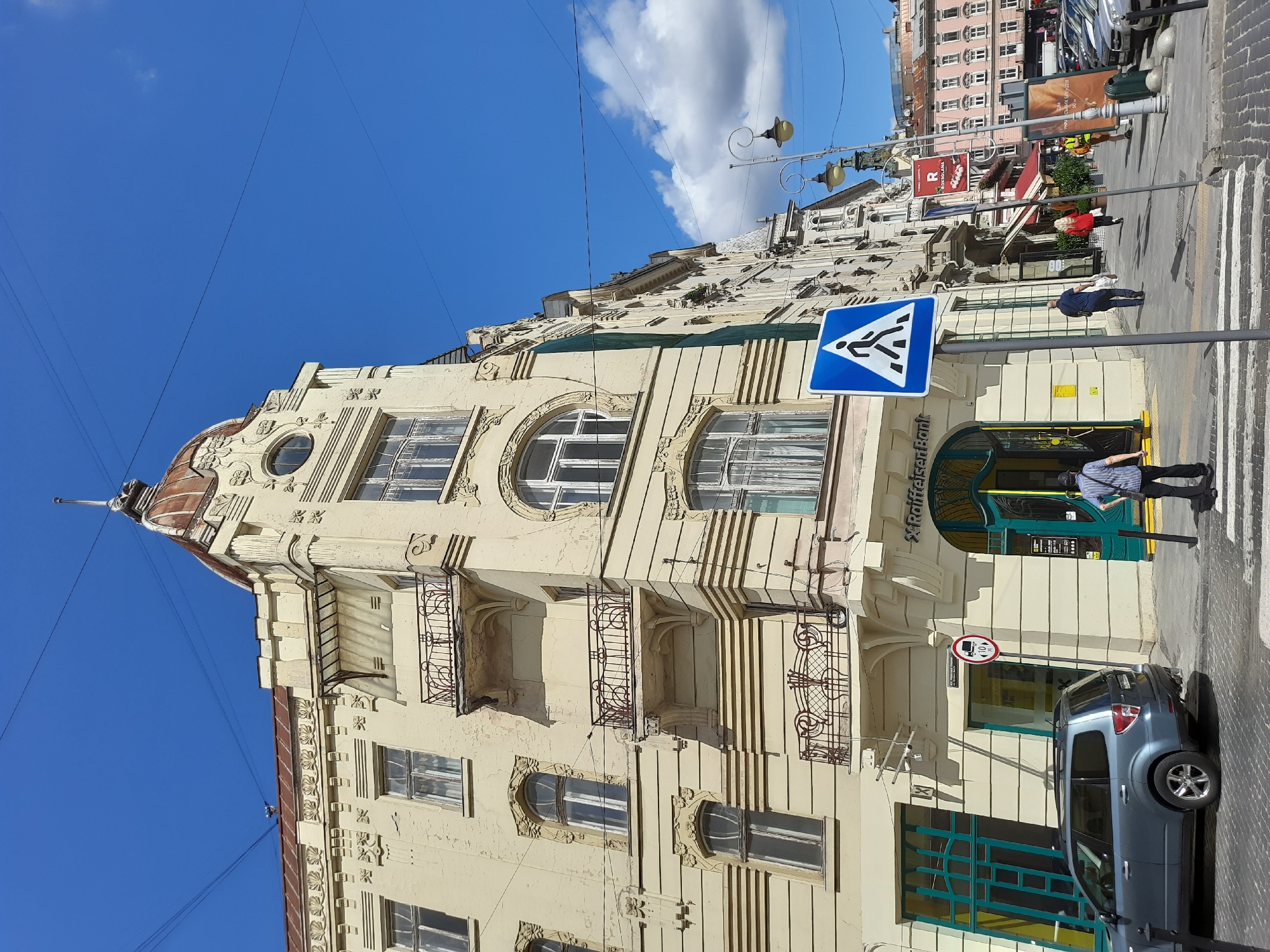 The image captures a street scene in Lviv, Ukraine, focusing on the facade of a cream-colored building with ornate architectural details. The building features multiple stories with arched windows, balconies with intricate metal railings, and a distinctive domed roof section. The ground floor appears to house a Raiffeisen Bank branch with a vibrant green entrance and signage.
In the foreground, a blue SUV is parked on a cobblestone street. A pedestrian crossing is visible, marked by white stripes on the road. A blue square sign with a white pedestrian icon indicates the crossing. A man wearing a striped shirt and dark pants stands on the sidewalk near the crossing, facing away from the camera. Further down the street, other pedestrians are walking.
The environment suggests a sunny day with a clear blue sky and scattered white clouds. Overhead, electrical wires crisscross the sky. The street is lined with buildings, some of which have awnings and signs. In the background, a variety of vehicles are parked along the street. The overall impression is a typical urban streetscape in Lviv.