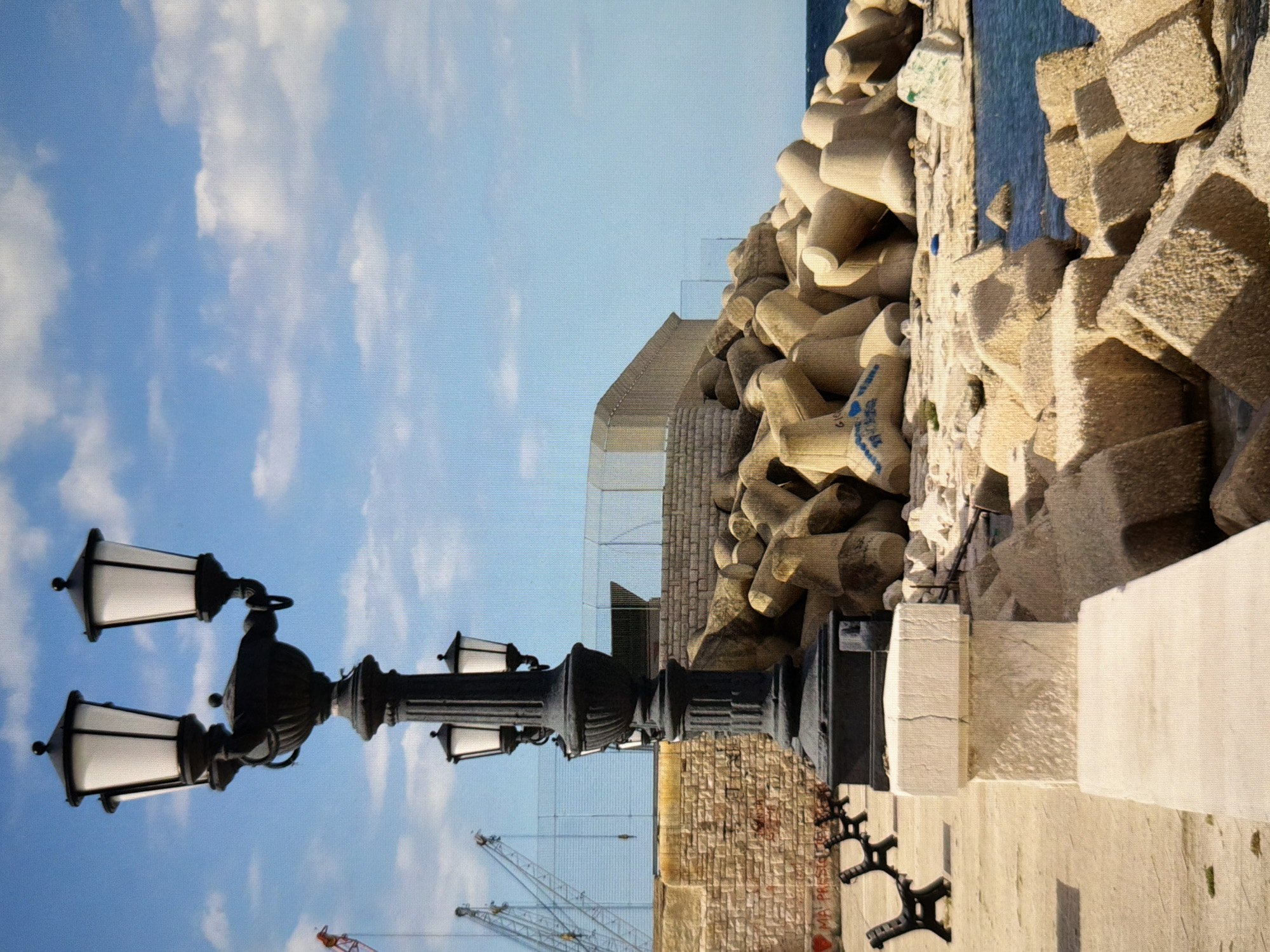 The image depicts a waterfront scene with a prominent, ornate black lamppost in the foreground. The lamppost has multiple lights and a decorative base. Beyond the lamppost, a paved walkway with several black benches lines the edge of the water. A stone wall with graffiti runs behind the walkway. The area is fortified with large, concrete tetrapods, piled along the shore to form a breakwater. In the background, a body of water is visible, meeting a bright blue sky with scattered white clouds. Industrial cranes are visible in the distance. The overall impression is one of a functional, perhaps industrial or port, area with public amenities. The scene does not contain any people or animals. The image is taken from a low angle, emphasizing the lamppost and the tetrapods. The lighting suggests it is daytime, with sunlight illuminating the scene. The location is identified as the city of Kyiv, Ukraine.