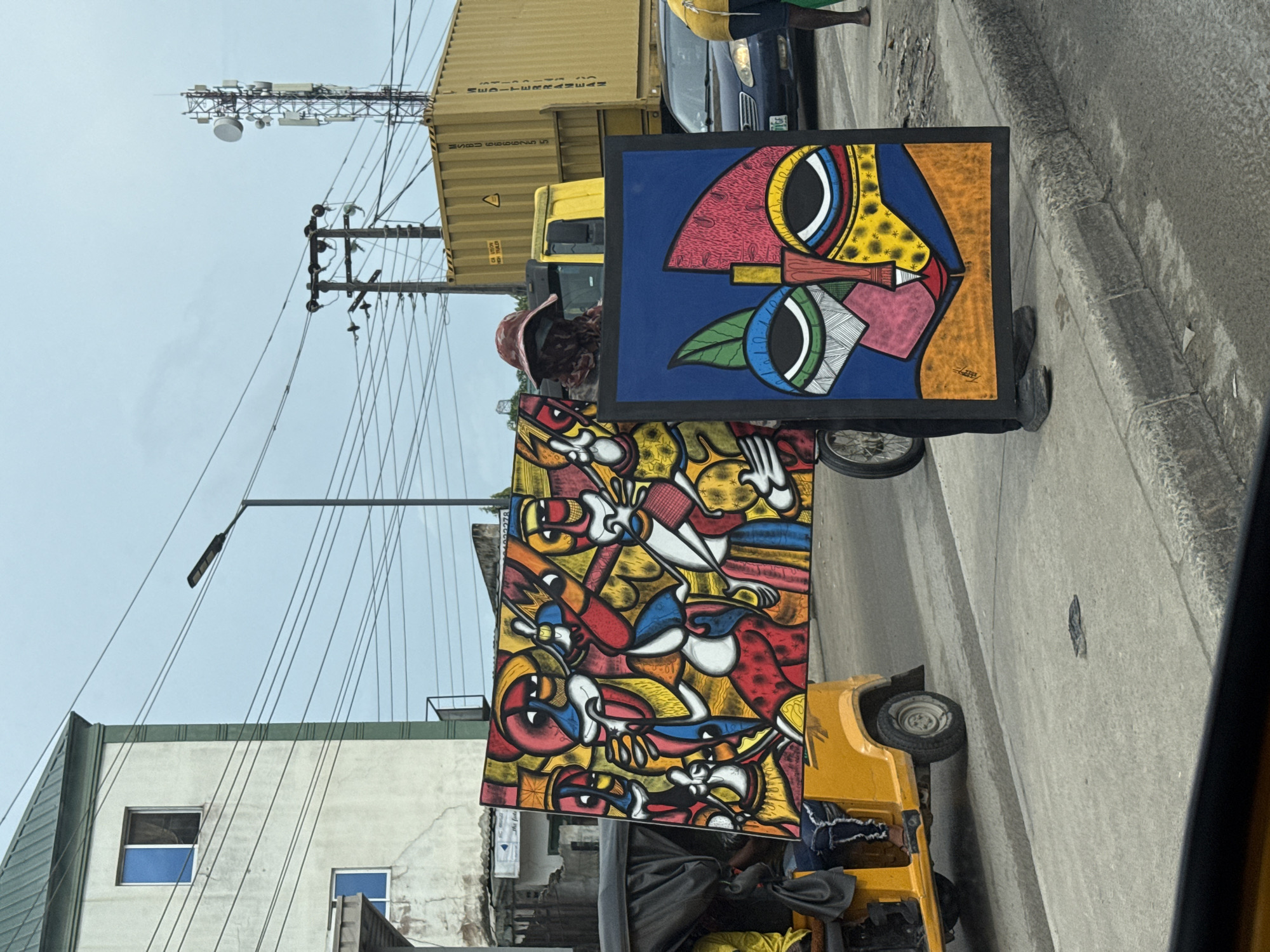 An outdoor street scene in Lagos, Nigeria, captures a vibrant display of abstract art being transported or displayed amidst urban activity. The image is oriented sideways, but depicts a bustling daytime environment under an overcast sky.
In the foreground, two large, brightly colored paintings are prominent. One artwork, positioned upright, features a stylized, cubist-like face in bold hues of red, yellow, blue, green, pink, and white, with distinct patterns and outlines. A smaller, illegible signature is visible on this painting. The second, larger painting, placed horizontally, presents a complex arrangement of multiple figures or abstract shapes, rendered in a similar vivid palette of red, yellow, blue, white, black, and orange.
These artworks are mounted on or beside a yellow tricycle, commonly known as a Keke Marwa or Napep in Nigeria, which appears to be carrying them. Parts of a person (or people) are visible near the tricycle, including legs and clothing, suggesting someone is operating or attending to it. Further down the street, a dark blue car is partially visible.
The background reveals typical urban infrastructure. A large yellow container truck, partially obscured, shows text like "MEDITERRANEAN" and the container number "USBU 666278," along with a caution symbol. Utility poles with a dense network of overhead wires crisscross the sky, alongside a street light. A cellular tower or antenna array is also visible against the light sky. Buildings with light-colored facades and windows, some with green trim, line the street. The ground consists of an asphalt road and a concrete sidewalk or curb. The scene suggests a lively, commercial area with a blend of transportation and artistic expression.