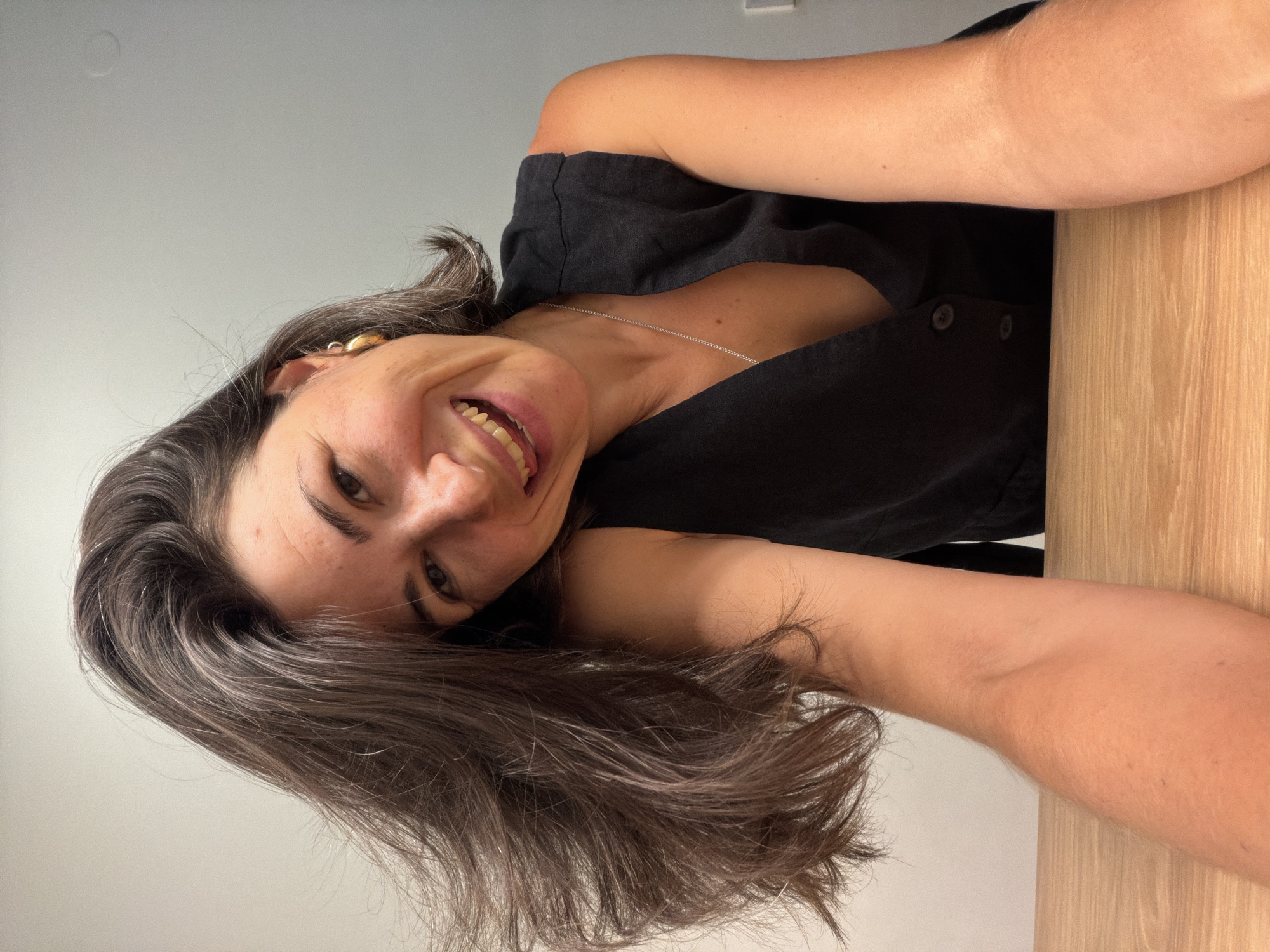 A woman with dark brown hair, streaked with some gray, is featured in a close-up, sideways portrait. She smiles broadly, revealing her teeth, and looks directly at the camera with a happy and engaged expression. Her dark eyes are visible, and her skin appears clear. She wears a black top or dress with a V-neckline and visible buttons down the front. A delicate silver chain necklace adorns her neck, and a prominent gold earring is visible in her right ear (left side of the image).
The setting appears to be indoors, against a light gray or off-white wall or ceiling. A light-colored wooden surface, possibly a desk or table, is visible on the right side of the frame. The lighting is bright and even, suggesting daytime. The overall mood of the image is cheerful and relaxed. The provided context states the location as Budva, Montenegro, though no specific geographical cues are visible within the image itself. There is no visible text.