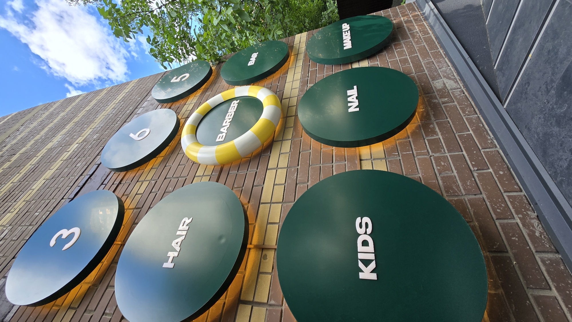 An eye-level, exterior, vertical medium shot captures a brick wall of a building in Kyiv, Ukraine, featuring a series of circular signs. The signs are an assortment of dark green and teal colored discs with white lettering. The top row of signs includes the number "3", "6", and "5". Below these, a yellow and white ringed sign reads "BARBER". Further down, dark green signs read "HAIR", "KIDS", "NAIL", and "MAKE UP". A few of the signs have orange lighting emanating from behind them. A tree with green foliage is visible on the right side of the frame, against a bright blue sky with scattered white clouds. The lower portion of the image shows a dark gray metal gutter or trim along the bottom of the brick wall, and a dark gray textured surface below that, possibly a sidewalk or walkway.