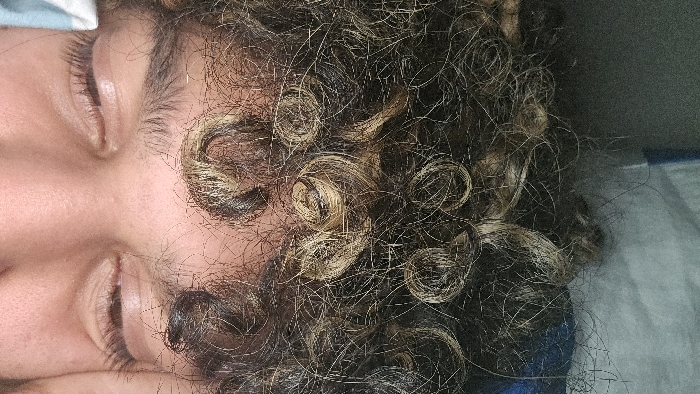 A person is lying on their side, eyes closed. Their hair is dark, curly, and highlighted with blonde streaks, forming tight ringlets around their face. Skin tone is fair. A blue fabric is visible behind the person's head. The image is shot from an overhead perspective, focusing on the person's face and hair.