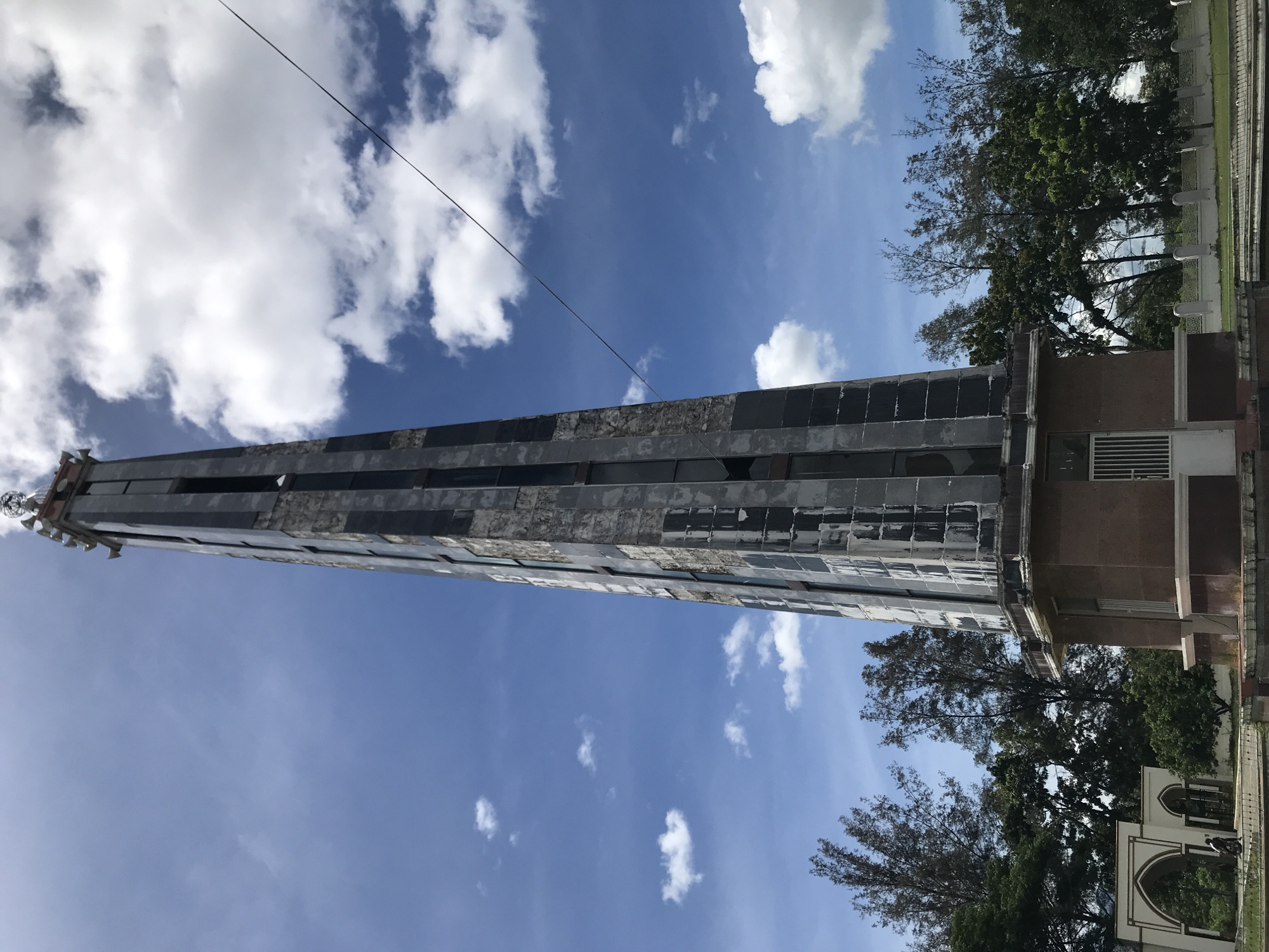 The image shows a tall, weathered monument composed of alternating light and dark stone blocks, situated in Jantho, Indonesia. The monument rises vertically against a backdrop of blue sky with scattered white clouds. A thin power line stretches across the upper part of the sky. The base of the monument is a broader, squarish structure with visible windows and a darker stone facade. Trees with green foliage stand to the right of the monument, partially obscuring a white arched structure in the background. The monument appears to be aged and potentially damaged, with signs of wear and tear on its surface. The overall scene suggests a historical or commemorative landmark within a natural or park-like environment.