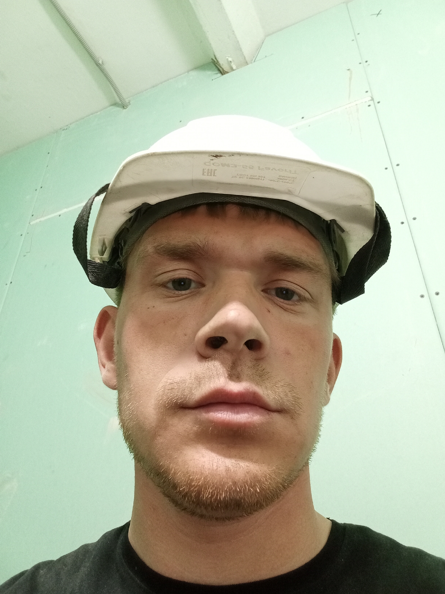 A close-up, low-angle, color photograph depicts a fair-skinned man wearing a white hard hat with a black strap. He has light brown hair and stubble. The background consists of a green wall made of drywall panels secured with screws. The setting appears to be an interior space under construction, possibly in a city with unknown details.