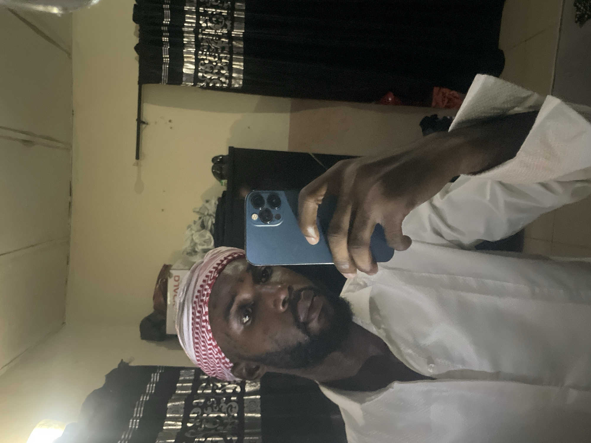 A man is taking a selfie in an interior setting. He is wearing a white shirt and a red-and-white patterned headscarf. He holds a blue smartphone in his hand. The background includes a dark curtain with a patterned design, a dark piece of furniture, and a light-colored wall. The location appears to be in a residential setting.