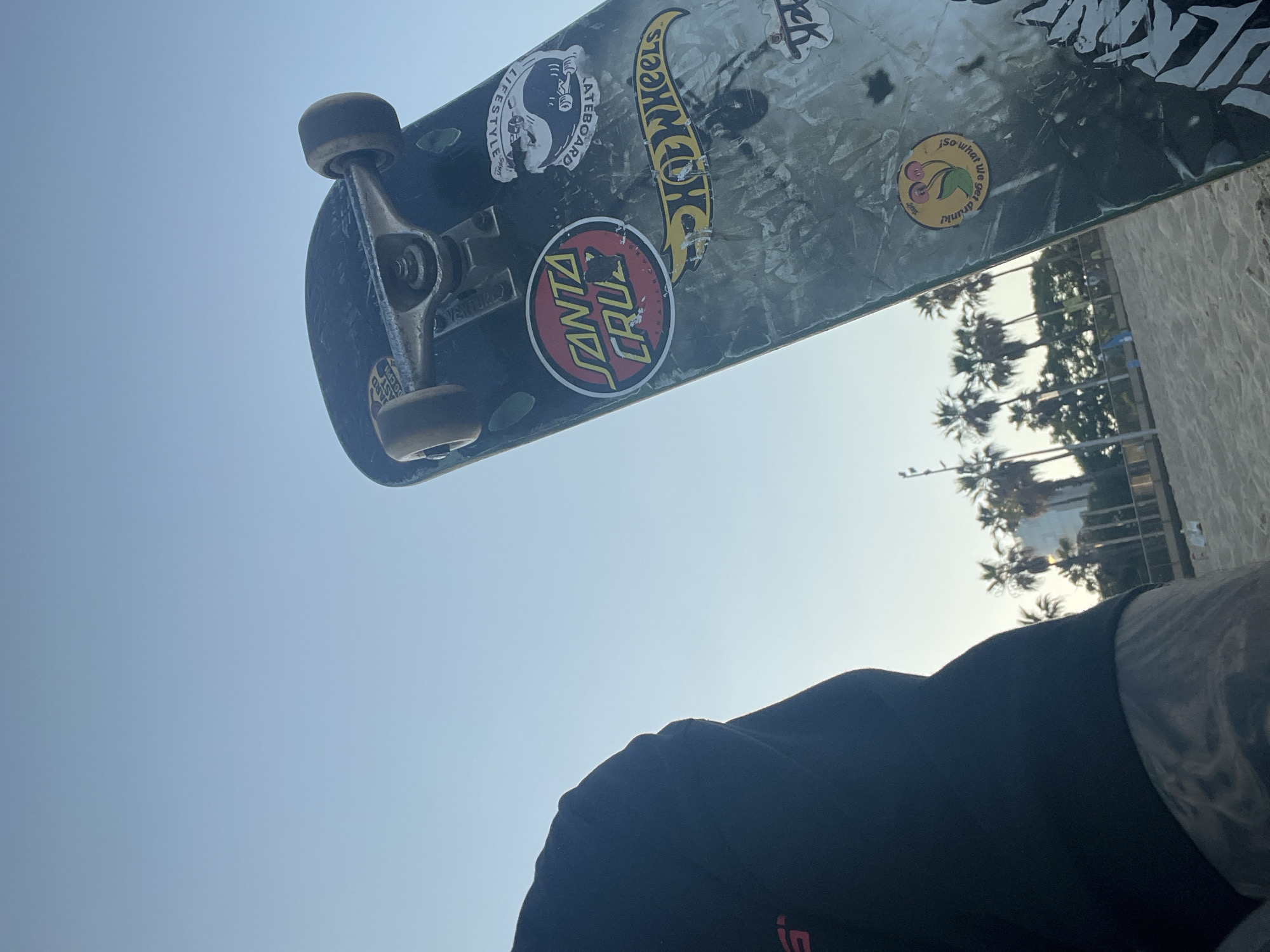 A low-angle shot captures a skateboard held in the air. Stickers adorn the underside of the skateboard, including "Santa Cruz" and "Hot Wheels" brands. In the background, palm trees suggest a coastal environment. The sky is light blue. A person's shoulder and neck, clad in a black shirt, are in the foreground. The scene does not have any details to place it in the city of Barcelona, Spain.