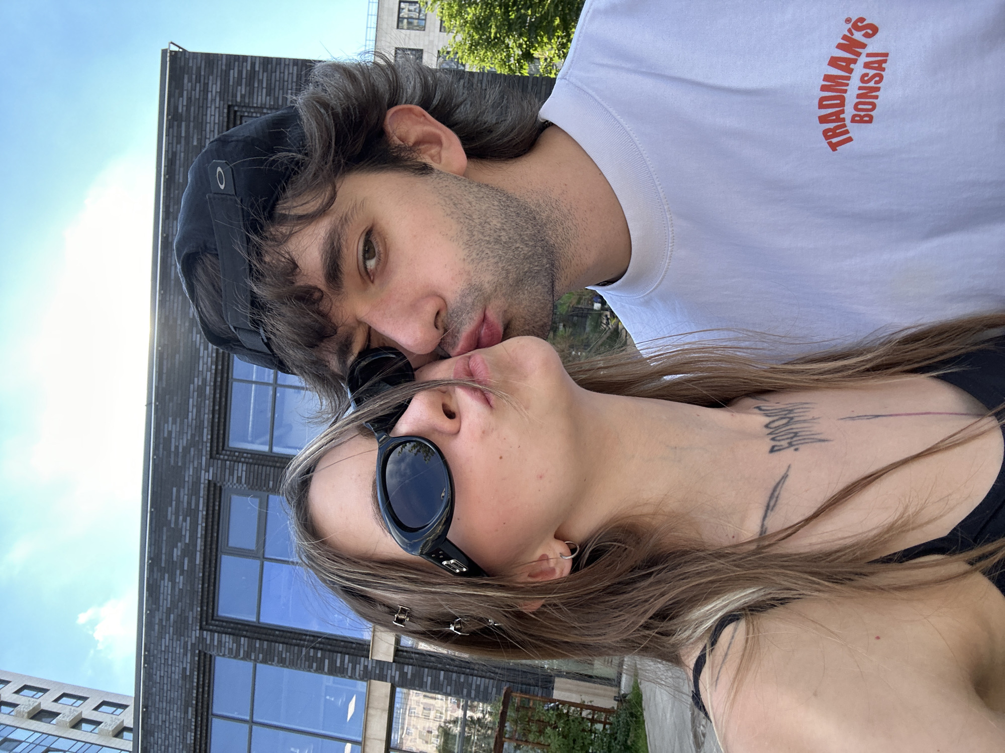 The media file shows a close-up selfie of a young couple in Moscow, Russia. The couple is outdoors, with a modern brick building and a blue sky visible in the background, suggesting it is daytime with good weather. The man is wearing a white t-shirt with red text that reads "TRADMAN'S BONSAI". He is also wearing a black baseball cap. The woman is wearing a black tank top and black sunglasses. Both individuals have their eyes closed, and their lips are touching in a kiss, indicating an affectionate moment. The woman has a tattoo on her chest and neck area. Small hair clips are visible in her hair. The overall mood is romantic and intimate.