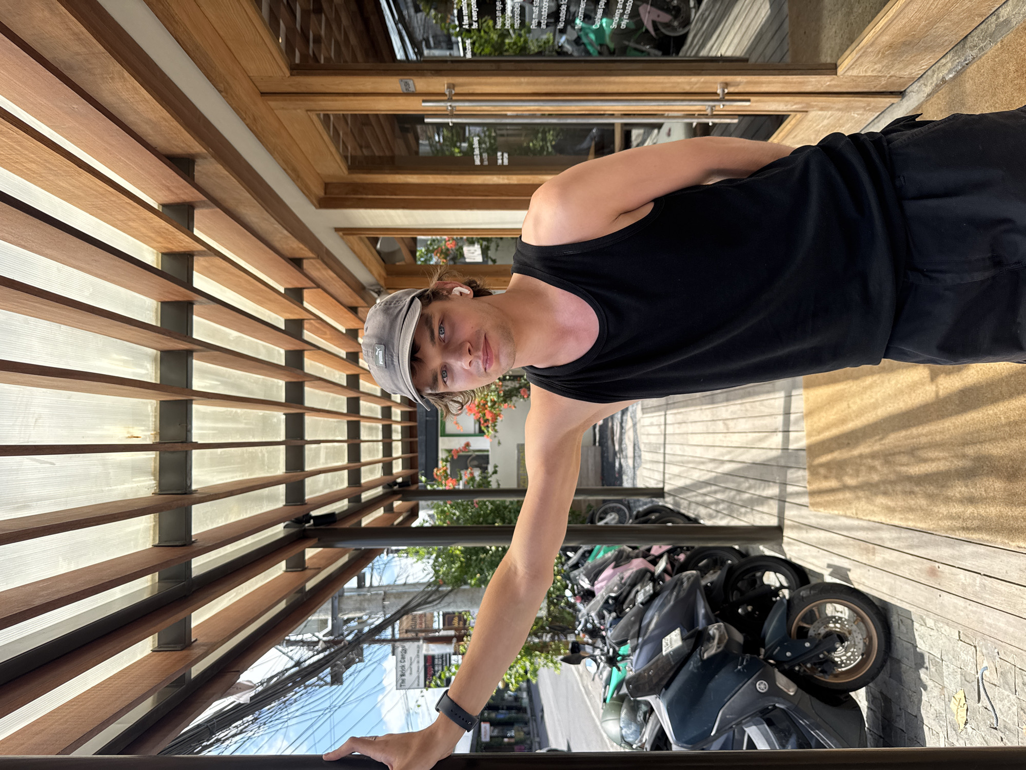 A young man in a black tank top and gray cap stands outside a building in North Kuta, Indonesia. He is leaning against a horizontal metal bar, holding it with his left hand which has a smart watch on the wrist. Behind him are parked motorbikes, and a sign on the wall indicates "The Brick Canggu" with some rules for patrons. The building's facade has a slatted wooden design.