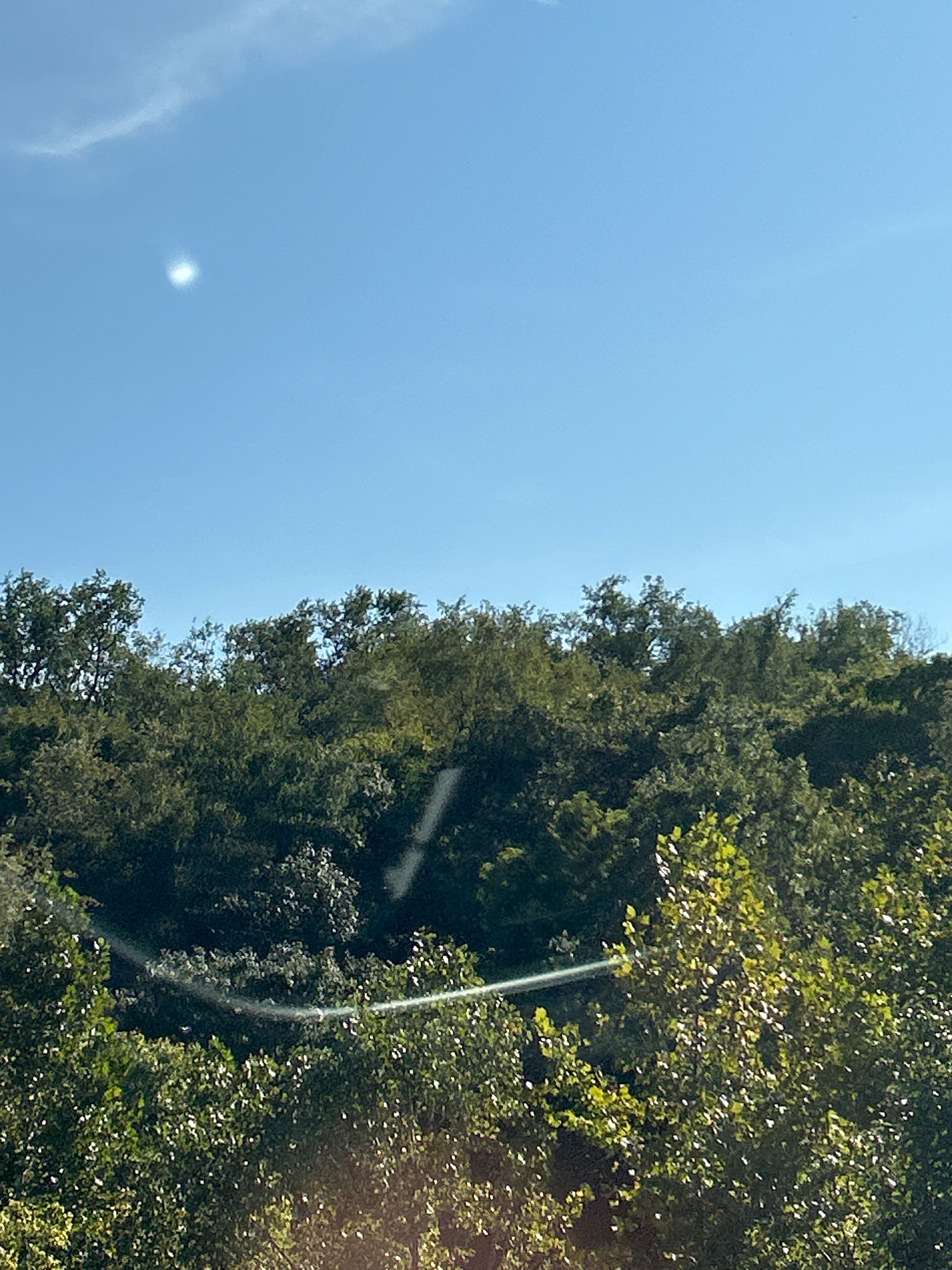 The image shows a clear blue sky with a bright sun, partially obscured by a thin cloud. Below the sky, a dense green forest covers a hill. In the foreground, the tops of trees are visible, with sunlight glinting off their leaves. There are no people or identifiable structures visible in the scene. The image appears to be a landscape photograph taken outdoors.