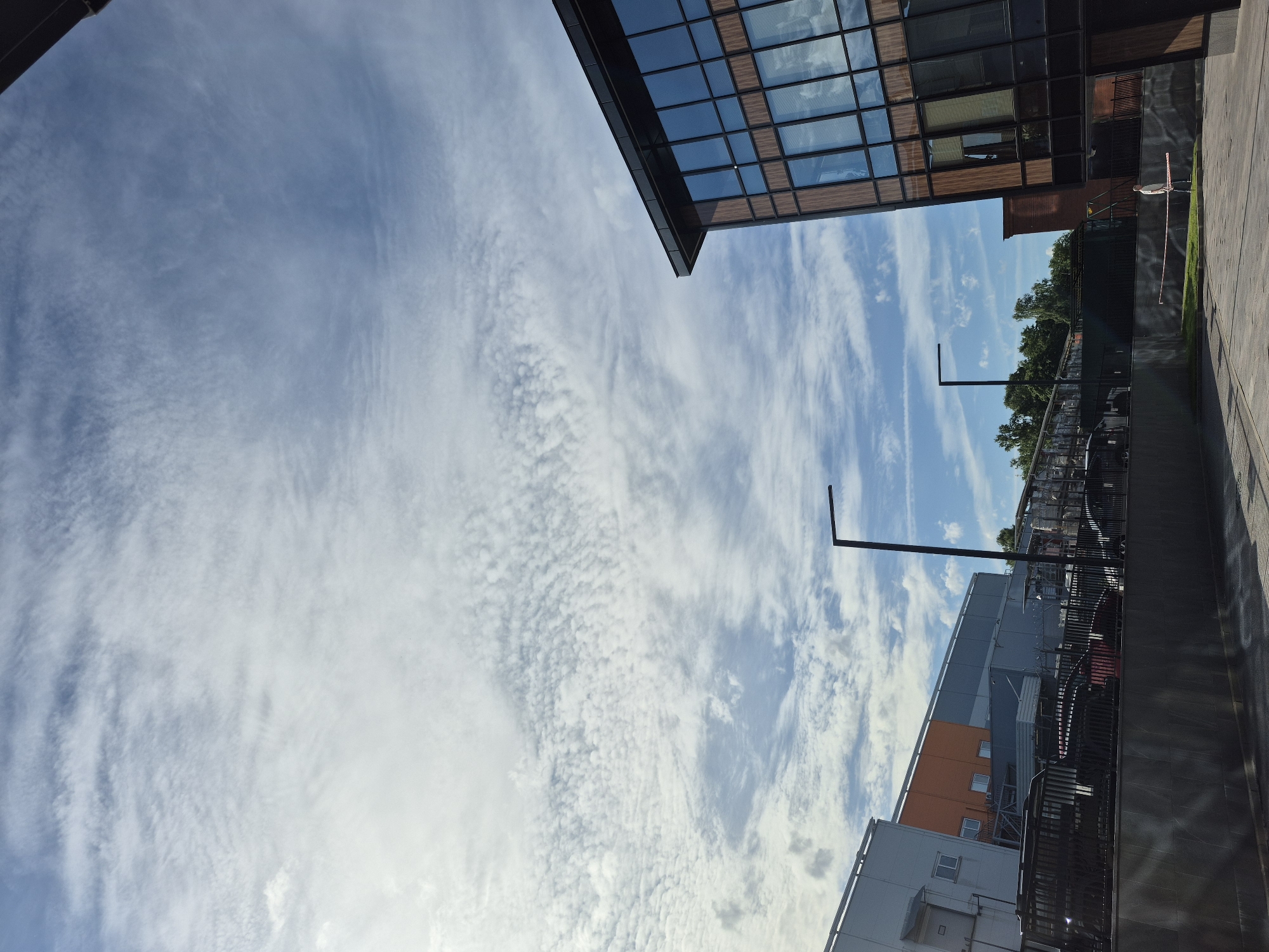 The image captures a view of modern buildings and a cloudy sky in what appears to be an urban setting, possibly Saint Petersburg, Russia. The perspective is from ground level looking upwards and slightly sideways.
The prominent subjects are the architecture of several buildings. One building in the upper part of the frame has a dark facade with large, reflective glass windows, suggesting a contemporary office or commercial space. Below and to the right, other buildings are visible, including one with orange and grey sections and another with a lighter grey exterior. These structures have multiple windows and visible infrastructure like railings and outdoor lighting fixtures.
The sky is a significant element, filled with a dynamic pattern of white, wispy clouds against a bright blue backdrop. The distribution of clouds suggests it could be late morning or early afternoon on a day with mixed weather conditions. Sunlight is evident, casting shadows and illuminating parts of the buildings and the ground.
There is a single person visible on the right side of the image, standing near a fence and a patch of grass. The person is dressed in light-colored clothing and is looking towards the side, away from the camera. A red and white striped pole is positioned near this individual.
The scene appears to be an outdoor walkway or plaza adjacent to the buildings, with paved ground and a metal fence. The overall atmosphere is neutral, focusing on the interplay of architecture, sky, and a hint of human presence. There is no discernible text within the image.