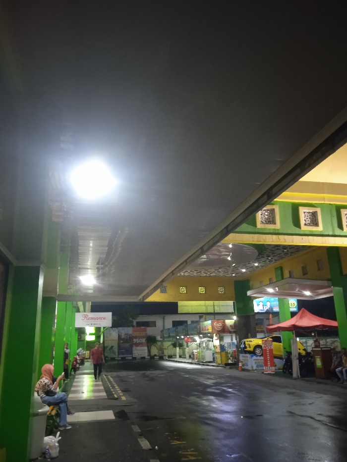 The image depicts a brightly lit gas station or rest stop at night in Kecamatan Kartasura, Indonesia. The ground appears wet, reflecting the numerous overhead lights, suggesting recent rain.
In the foreground on the left, a woman wearing a hijab and a patterned top sits on a white bench, engrossed in her phone. Further ahead, a man in a red shirt walks across a marked pedestrian area. The left side of the scene is framed by tall green pillars supporting a large, flat gray canopy that extends across the top of the frame.
The station's structure to the right features a distinctive design with green pillars and a yellow fascia, complemented by decorative patterns on the ceiling. Several fuel dispensers are visible in the mid-ground. A prominent display features a bright yellow car elevated on a platform, situated next to a red tent or canopy. Various other vehicles, including motorcycles, are parked around the facility.
Several visible signs and texts include "Romance" on a white sign on the left, "950K" on an advertising banner, and a green sign with "3.5" near the yellow car display. Other advertising banners with unreadable text are also present. Multiple individuals are scattered throughout the scene, either walking or sitting, indicating an active, though not bustling, environment. The overall impression is that of a functional and well-illuminated roadside facility during the evening hours.
