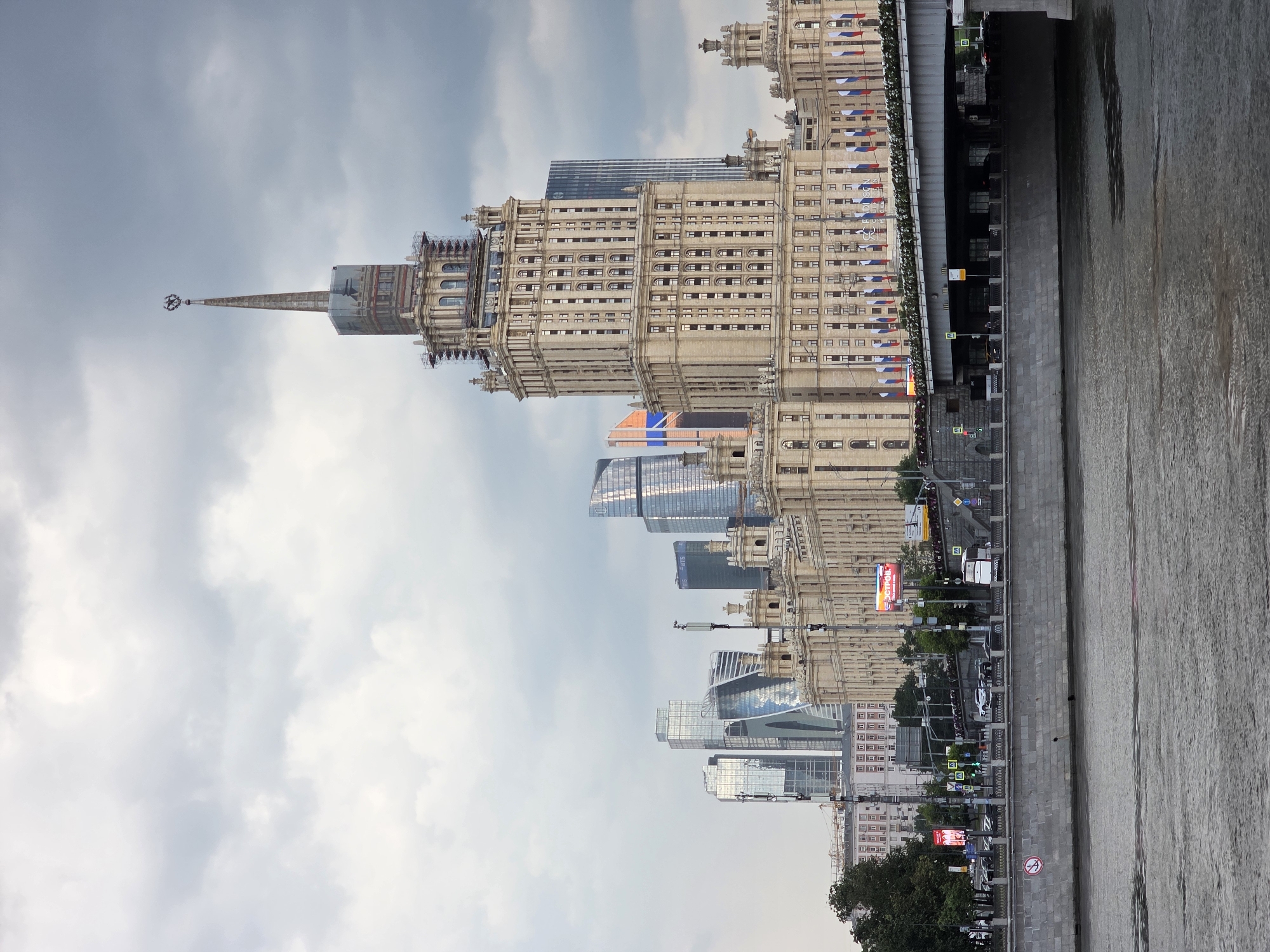 The image depicts a view of the Moskva River in Moscow, Russia, with the iconic Stalinist skyscraper, the Hotel Ukraina (now the Radisson Collection Hotel), prominently featured on the right side of the frame. The historic hotel, with its distinctive spire topped by a star, is adorned with scaffolding, suggesting ongoing renovation work. In the background, modern glass skyscrapers of the Moscow City business district rise into the cloudy sky. A bridge crosses the river in the foreground, with cars and a bus visible on it. The river's surface is calm, reflecting the overcast sky. The scene is captured from a slightly elevated vantage point across the river, offering a panoramic perspective of Moscow's architectural contrast between its historic and contemporary structures.