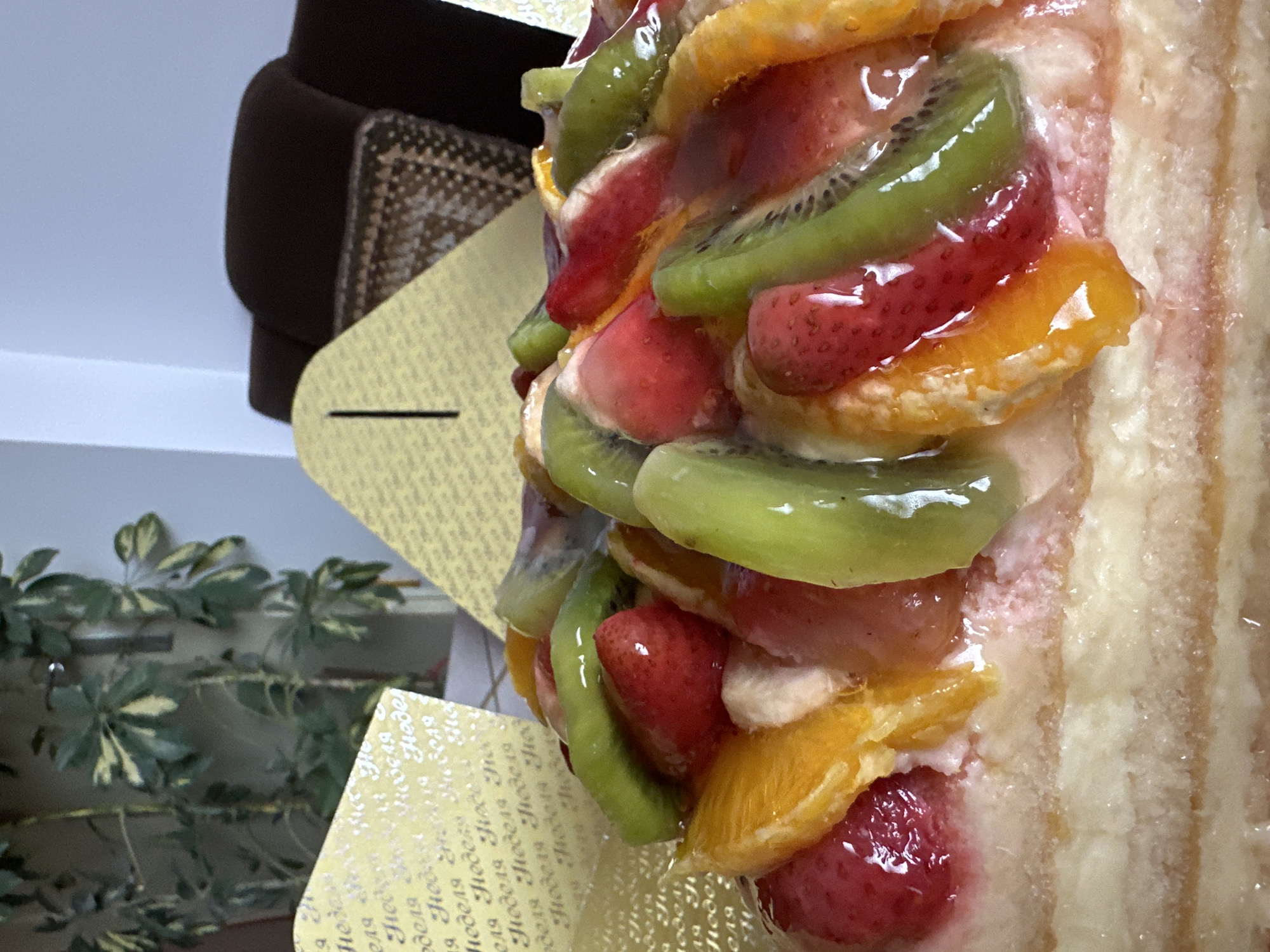 The image is a close-up of a fruit cake with visible layers of cake and a topping consisting of glazed slices of kiwi, strawberries, and oranges. A card with repeating text "Неделя" (Week) is partially visible behind the cake. The background includes a plant and a piece of furniture, suggesting an indoor setting. The location in the city of Montana, Bulgaria cannot be determined from the given visual information.