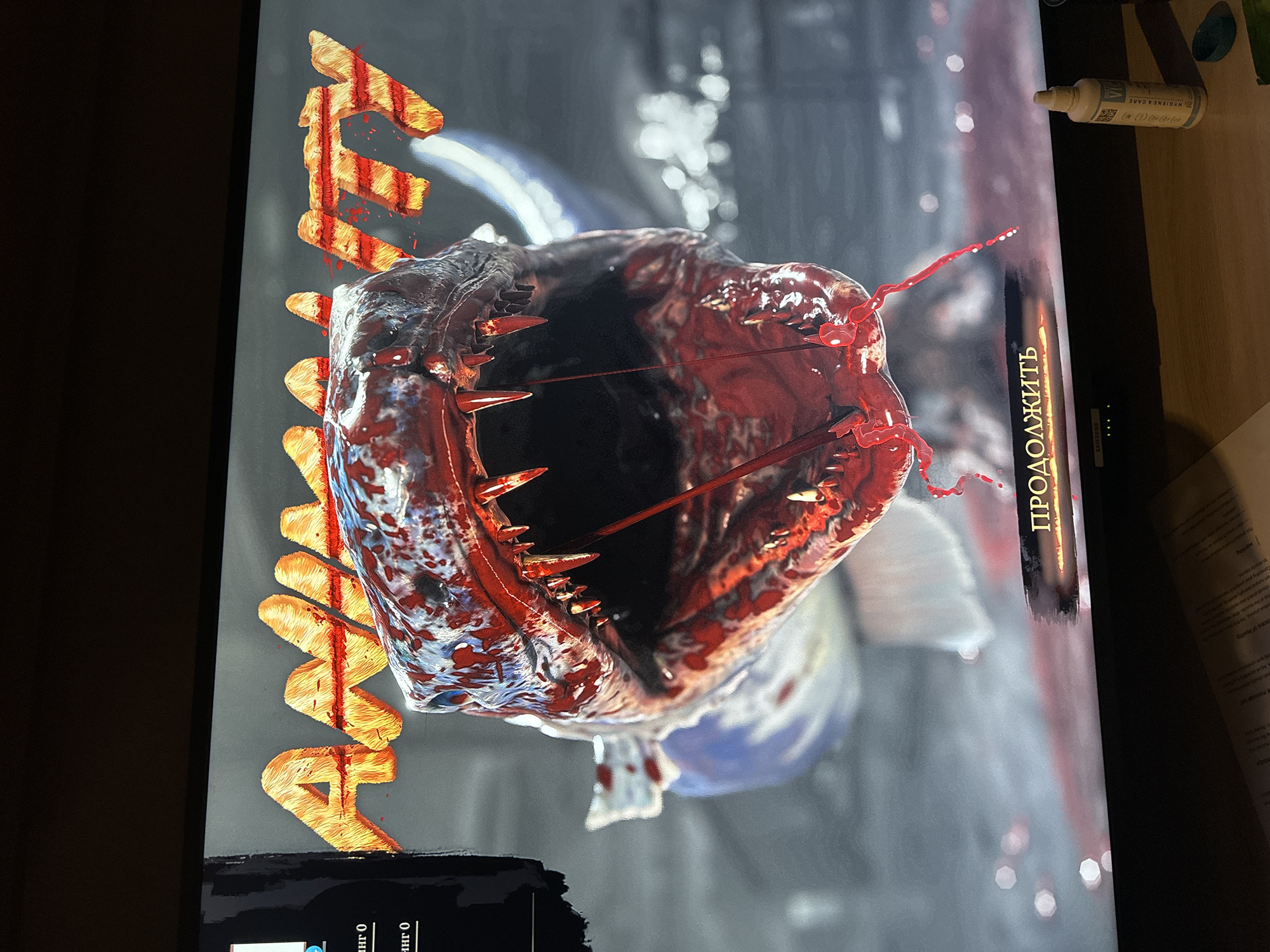 The image depicts a close-up view of a digital screen displaying a gruesome scene, likely from a video game or graphic design project. The primary subject is a monstrous creature's open mouth, heavily rendered with blood and sharp teeth. The word "AMA" is stylized and positioned beside the creature, possibly serving as a title or identifier.
The environment surrounding the screen includes a wooden surface, which suggests the scene is indoors. The city of Magnitogorsk, Russia, is not explicitly present in the image and no elements suggest this location. Other details on the wooden surface include hygiene products and papers.
The word "ПРОДОЛЖИТЬ" (which translates to "CONTINUE") in Cyrillic script is written vertically to the right of the monstrous mouth.