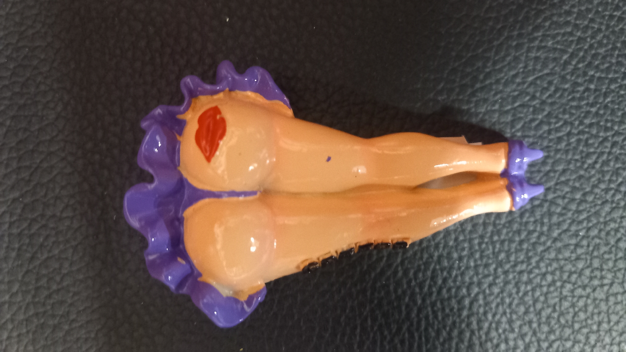 A novelty item resembling legs with a purple ruffled skirt and red lips painted on the buttocks is placed on a textured black surface. The item is positioned in the center of the frame, with the legs extending downwards and the skirt upwards.
