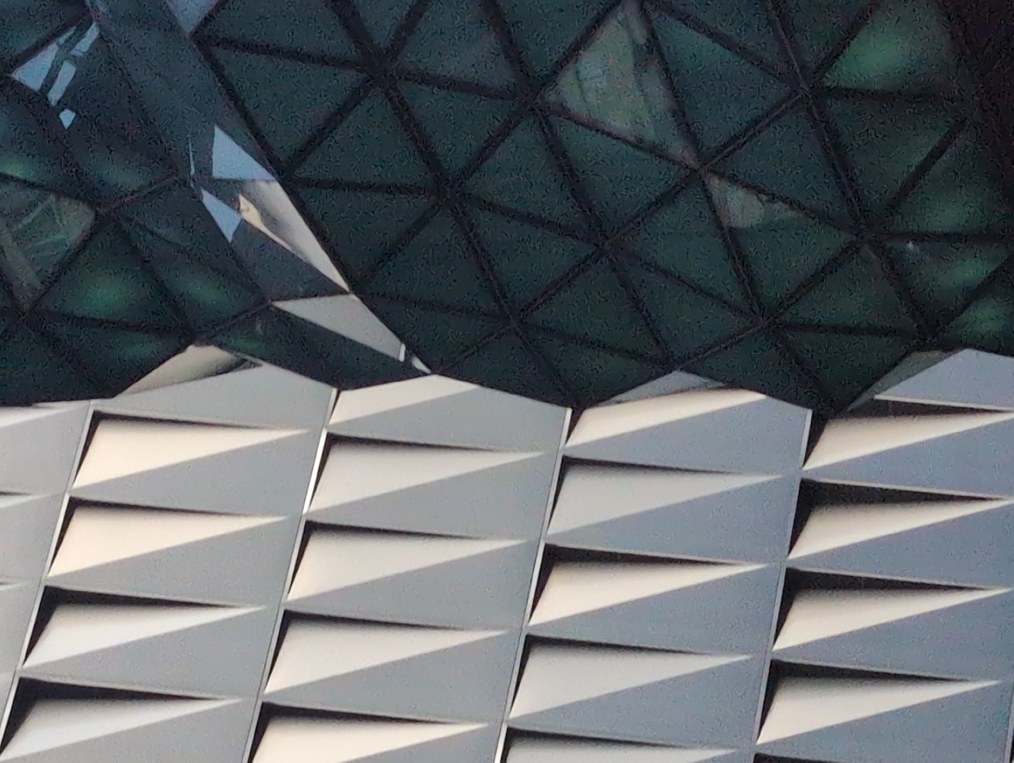 The image shows a close-up of two contrasting architectural facades. On the left, a grey, geometric facade features a repeating pattern of triangular indentations, creating a textured, almost scale-like appearance. The indentations are sharply defined, with strong contrasts between light and shadow. On the right, a dark, faceted glass facade dominates the frame. This section appears to be part of a curved building, with the glass panels forming a complex, diamond-shaped grid. The glass has a deep green, almost black hue, and reflects hints of the sky or surrounding structures, appearing somewhat mottled and textured. The two facades meet diagonally across the center of the image, forming a stark visual juxtaposition of matte and reflective surfaces, and angular versus curved forms. The image does not depict any people or specific environmental details that would allow for identification of a location within Kyiv, Ukraine.