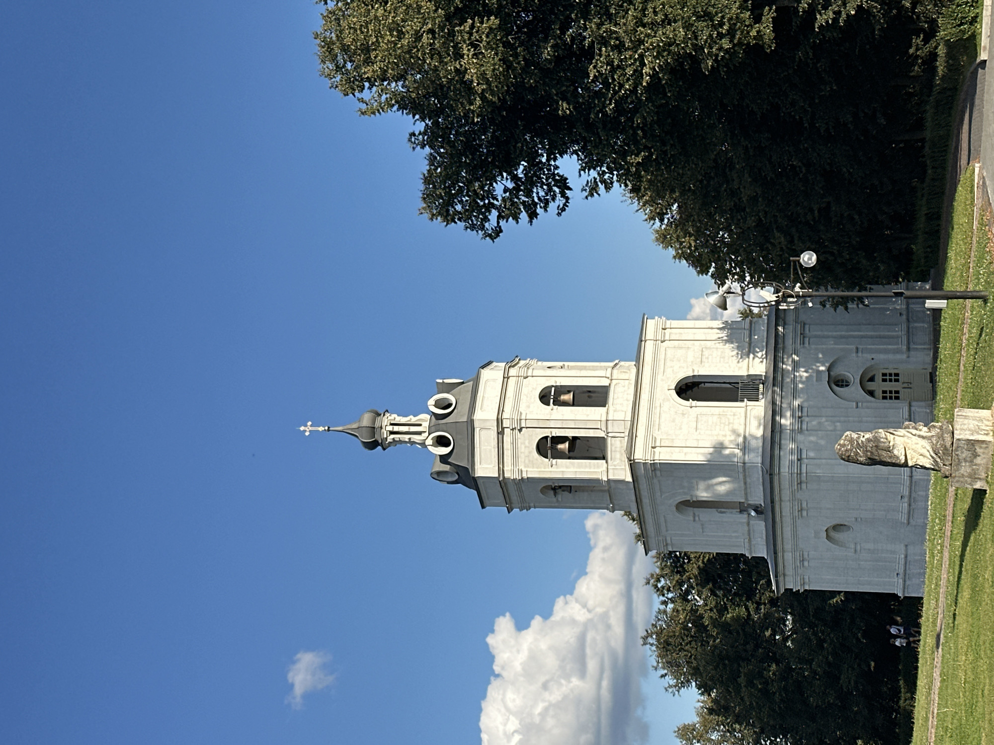 A white stone bell tower with a dark roof and a cross on top stands in the foreground. Bells are visible in arched openings. The tower is surrounded by lush green trees and a clear blue sky with scattered white clouds. In the lower left of the image, two people are walking on a grassy path. A stone sculpture is in the middle of the frame, and a lamppost with speakers is to the right. This is the Church of the Icon of the Mother of God in Dubrovitsy, Russia.