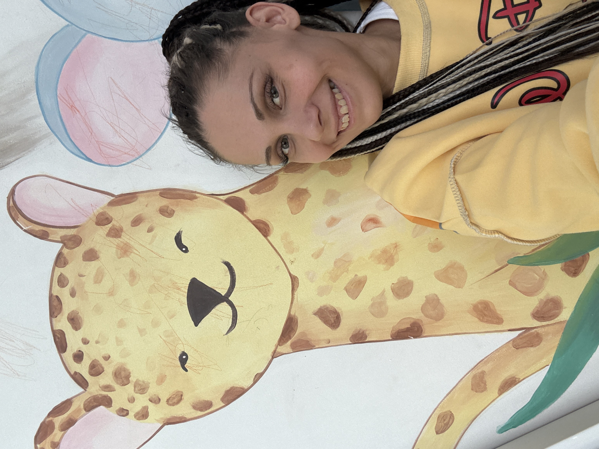 A woman with braided hair is smiling and posing in front of a mural. The mural features a cartoon cheetah lying down and two balloons above the cheetah's head, one pink and one blue. The woman is wearing a yellow sweatshirt. The location is Rusanovo, Russia.