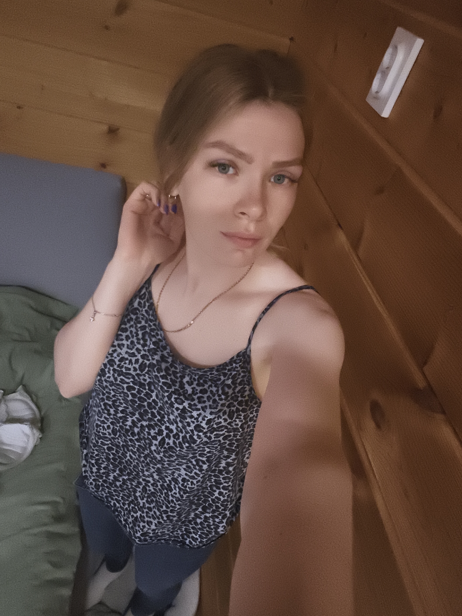 A woman is in a room constructed of wooden planks. She is wearing a dark tank top with a speckled pattern. She is also wearing blue pants and white socks. She is holding her hand up to her ear. There is a bed or couch behind her with a gray pillow and a green cushion. There is a small white object affixed to the wooden wall. I cannot confirm whether the location is in the city of Русский Вожой, Russia from the image provided.
