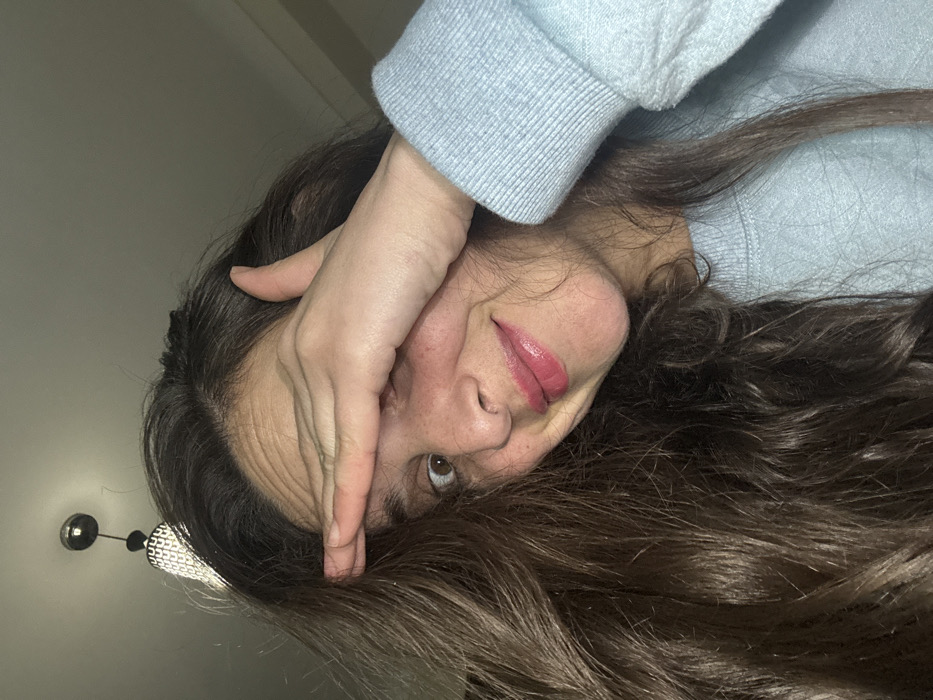 A person lies on their side with their hand covering the upper part of their face. The visible eye peers upward. The person has long, dark brown hair that flows across the lower half of the image. They wear a light blue sweater with a ribbed cuff. Their lips are coated in pink lip gloss. A decorative light fixture is visible on the upper left.