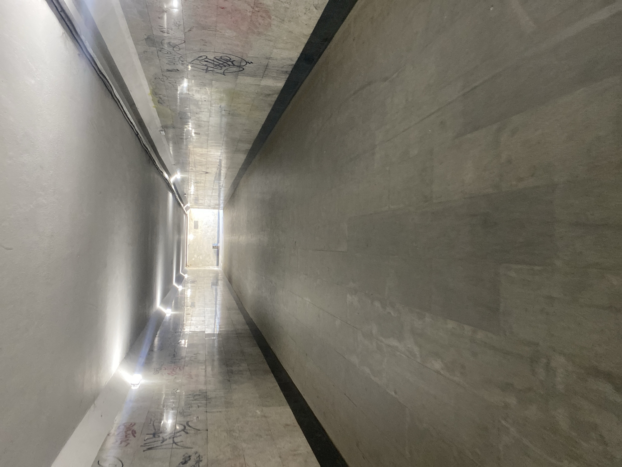 A long, narrow, empty corridor or passageway is depicted, viewed from a slightly elevated perspective looking straight down its length. The space appears to be constructed of concrete and features distinct material differences on its opposing walls and floor.
The left wall is smooth, light-colored (possibly white or light grey), and illuminated by a series of small, bright, upward-casting light fixtures mounted low on the wall. These lights create strong beams and reflections on the highly polished or wet-looking floor adjacent to this wall. A thin, dark line, possibly a cable, runs along the upper edge of this wall where it meets the ceiling.
The right wall is darker, textured, and appears to be made of rough concrete or stone panels. It is less reflective than the left wall and floor.
The ceiling is concrete, showing faint, scattered graffiti tags and reflections from the floor lights. The floor itself is highly reflective, particularly on the left side, mirroring the lights and ceiling. Extensive, dark graffiti is also visible on the left side of the floor. A distinct, narrow, dark channel or strip runs longitudinally down the center of the floor, creating a separation between the brighter, reflective left side and the darker, less reflective right side.
A bright, rectangular opening at the distant end of the corridor suggests an exit or entrance to another illuminated area. No people or specific activities are visible. The graffiti on the ceiling and floor is largely illegible, consisting of various tags and shapes. The overall impression is one of starkness and depth, with the artificial lighting creating a leading line towards the distant light source.