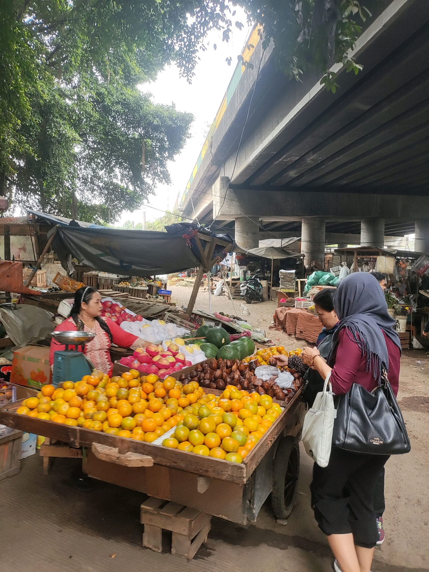 In East Jakarta, Indonesia, beneath an elevated highway structure, an outdoor market scene unfolds. A woman wearing a red and white patterned top and a black headband, with her hair pulled back, stands behind a wooden fruit cart laden with oranges. To her right, a scale with a metal bowl sits on the cart. The cart itself is filled with numerous oranges, with some watermelons and other fruits like rambutan visible in the background. A woman wearing a dark purple hijab and a maroon shirt, holding two bags, one white and one black, interacts with another woman who is partially visible. The market stalls are covered with dark tarps. The environment suggests a busy, open-air market typical of the region.