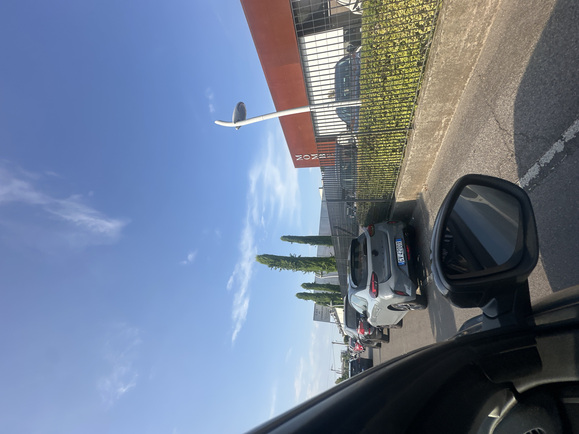 The image, taken from inside a vehicle, shows a traffic jam on a sunny day in Chiari, Italy. A line of cars is backed up on a street lined with trees and buildings. One building, visible in the background, has "MOMBE" in large letters on its facade, and another sign reads "CASA COMMUTATO". A black car is in the foreground, and the rear of a grey hatchback with license plate GV 420DX is visible. The sky is blue with a few wispy clouds.
