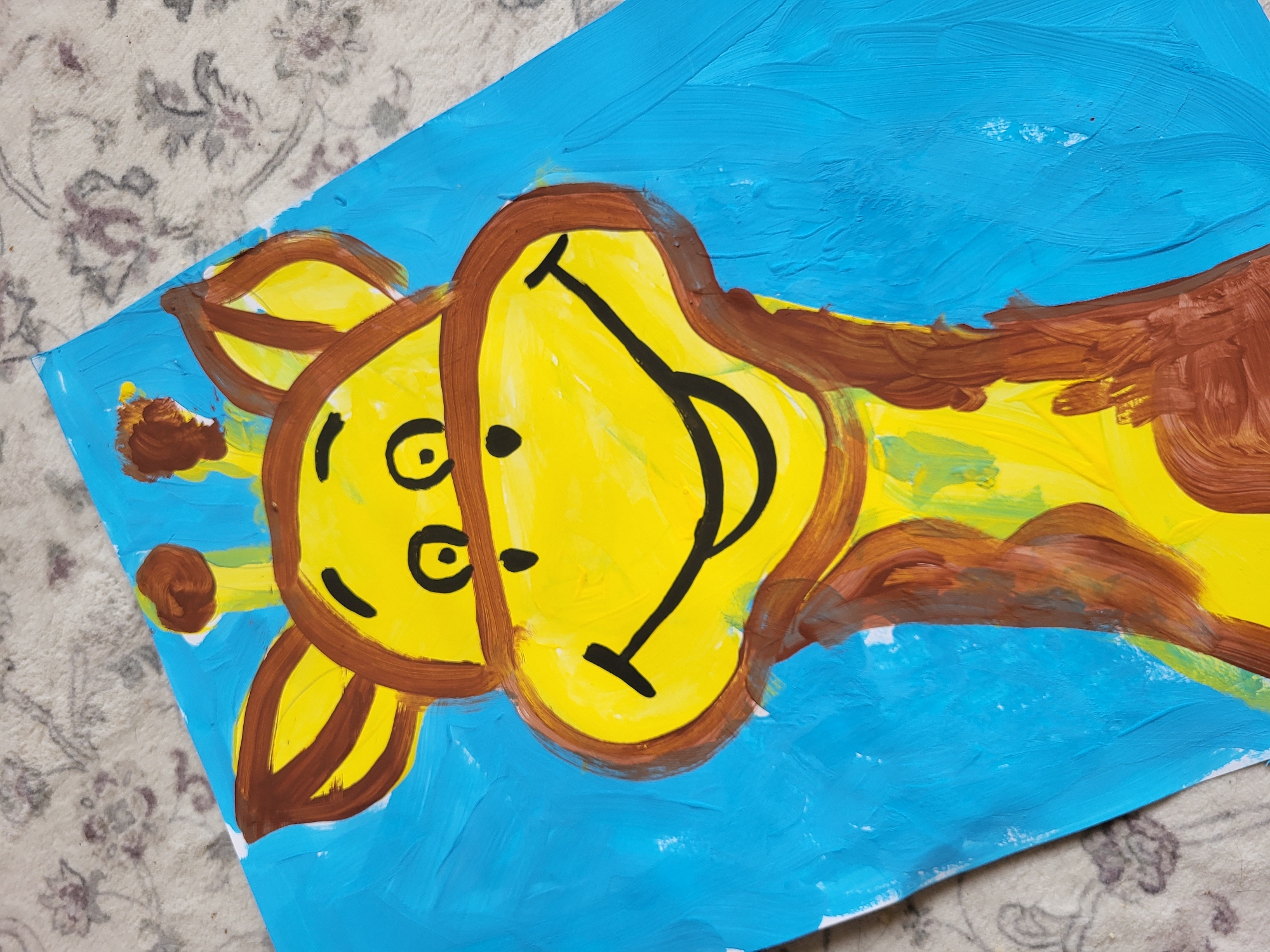 A close-up, high-angle shot features a brightly colored painting of a cartoon giraffe's head and neck on a blue background. The giraffe has a large yellow face with big, round eyes and a wide, black smile. Brown paint is used for the giraffe's ears, horns, and spots, as well as to outline its face and neck. The background is a solid, textured blue. The painting is resting on a surface with a floral pattern. The image is cropped, so the full extent of the painting and its surroundings are not visible. There are no people or any other identifiable objects in the image besides the painting and the patterned surface it lies on. The location is unknown.