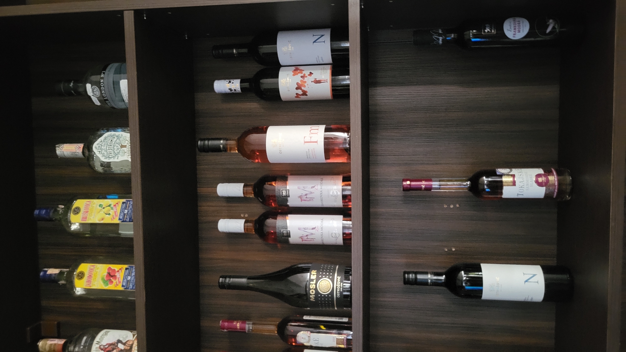 A tiered shelf unit made of dark wood displays various bottles of alcohol. The top shelf holds four bottles: a clear bottle with a red and gold label, a yellow-labeled bottle, a green-labeled bottle, and a clear, decorative bottle with a light-colored label. The middle shelf features six bottles: a dark brown bottle with a gold and black label, a clear bottle with a black label, two clear bottles with white labels and a pinkish liquid, a clear bottle with a pink liquid and a white label, and a clear bottle with a black label. The bottom shelf contains four bottles: a clear bottle with a black label, a clear bottle with a pinkish-red liquid and a white label, and a clear bottle with a dark label. The background is a dark wooden surface. The scene appears to be an indoor setting, possibly a bar or a private collection. The specific location within Prešov, Slovakia, cannot be determined from the image alone.