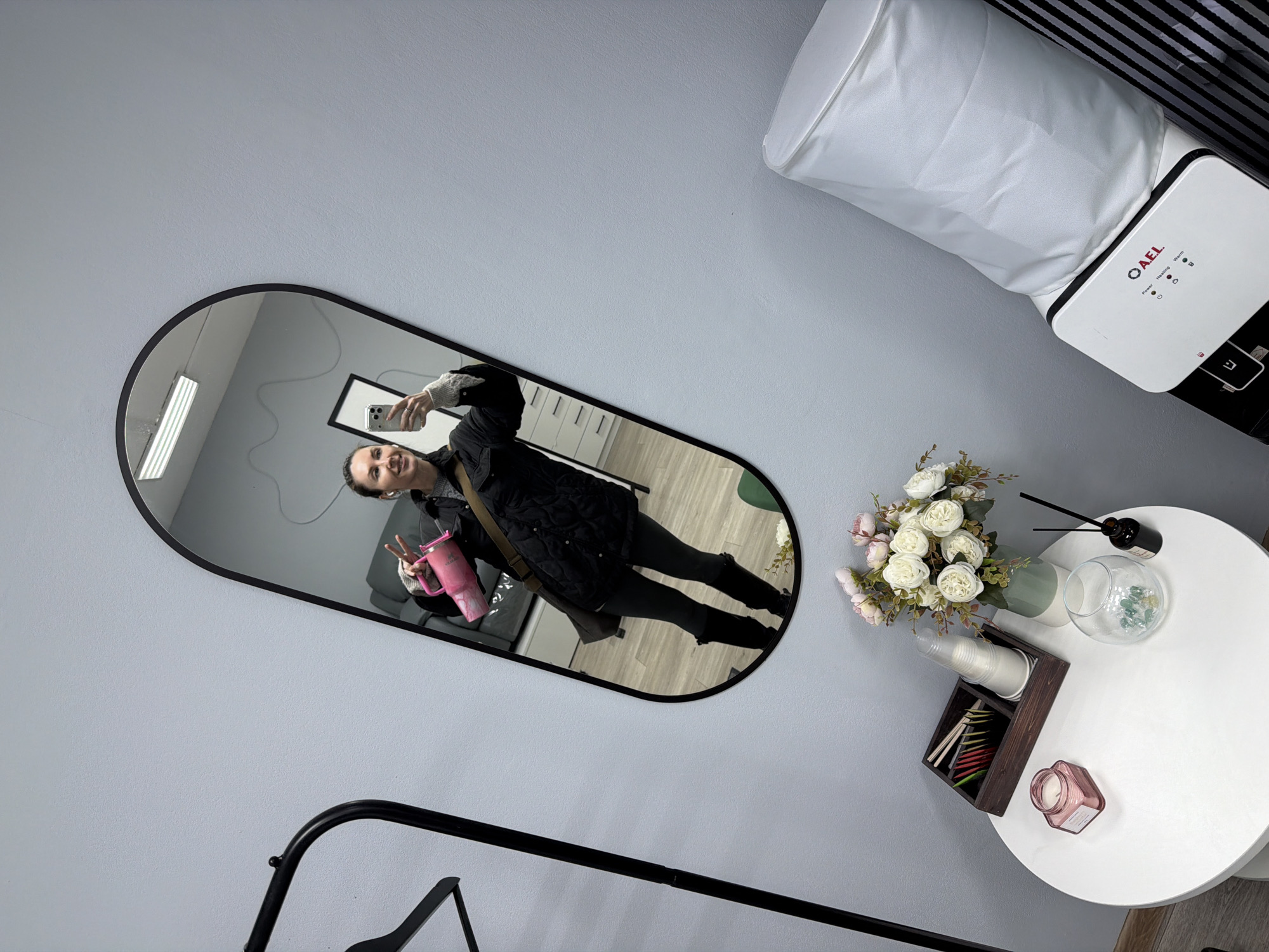 A person takes a selfie in a mirror in a room in Perm, Russia. The mirror reflects a woman wearing a black jacket, leggings, and boots, holding a pink tumbler and making a peace sign. The room features a round white table with flowers, tea, and a diffuser, and a piece of equipment with the letters "AEL". A clothing rack is also visible.