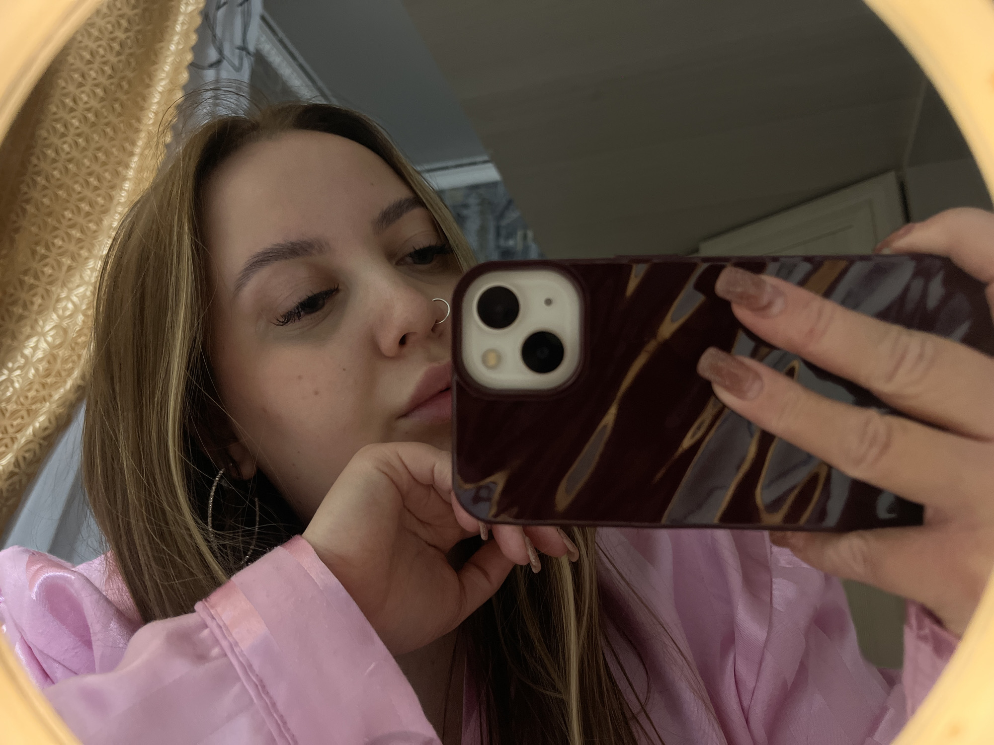 A woman in a pink robe is taking a selfie in a mirror. She is holding a phone in front of her face, with the camera visible. She has blonde hair, a nose ring, and is wearing earrings. The background is partially visible, showing a wall and a curtain. The location is indicated to be Novopokrovskaya, Russia, but there is no specific evidence of this in the image.