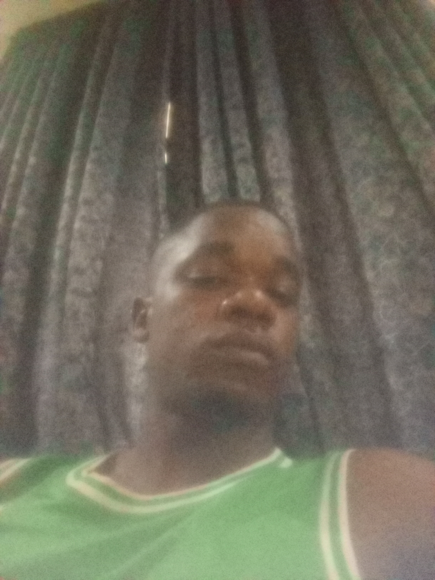 A close-up, blurry image depicts a man wearing a green and white trimmed tank top. He appears to be indoors, with what seems to be patterned curtains visible behind him. The location is Unknown, Unknown.