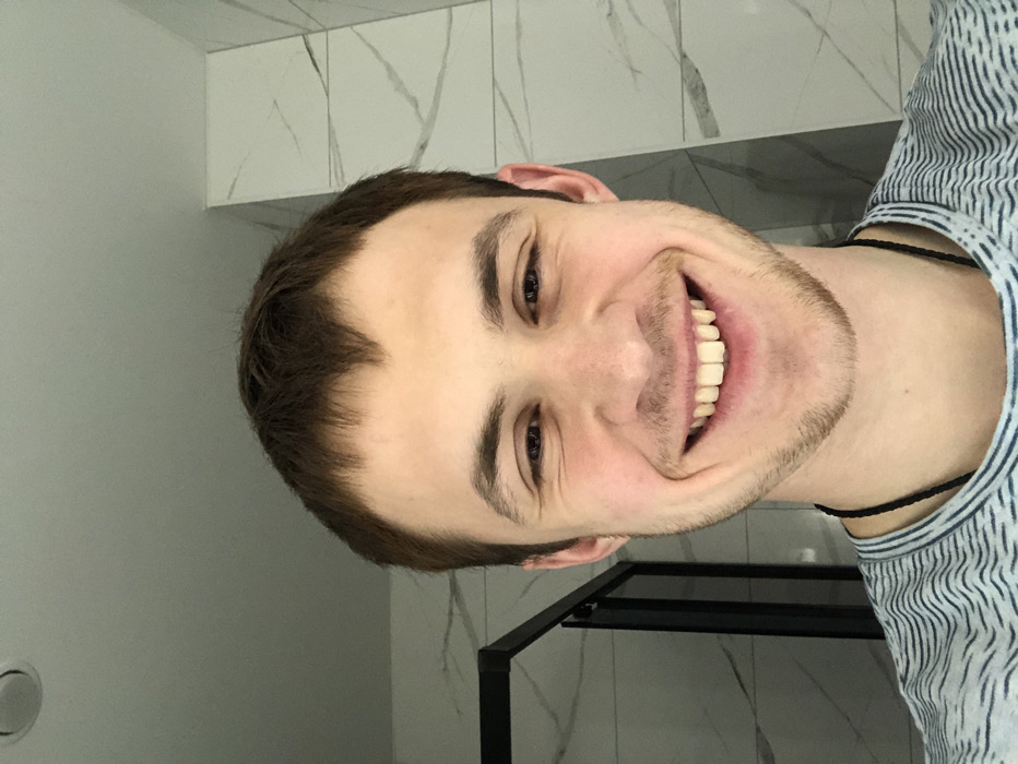 A smiling man is oriented sideways in a bathroom-like setting with gray tile. He has short, dark hair, light skin, and visible stubble. He wears a light-colored, striped shirt and a thin, dark necklace. A black metal frame is visible behind his shoulder. The room features white cabinets or tiling with gray marbling. A light gray wall is seen in the background, with a round, light-colored fixture on it.
