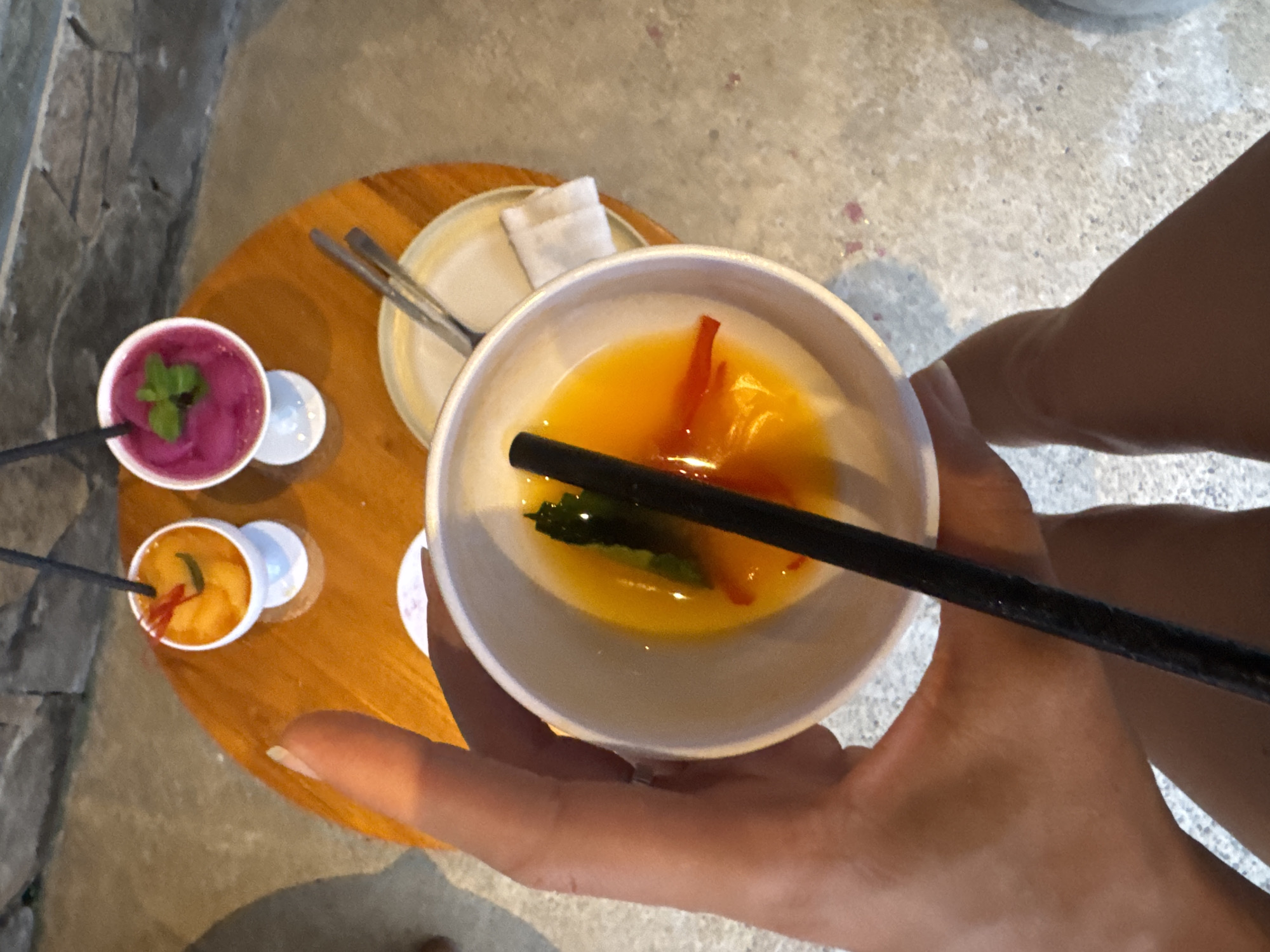 The image provides an overhead view of a person holding a small white bowl containing a yellow liquid garnished with pieces of red and green, possibly fruit or herbs. A black straw or chopstick rests in the liquid. On a round wooden table, two other small servings are visible: one with a purple-colored liquid topped with mint leaves, and another with an orange liquid, both in white cups with black straws.
Adjacent to the purple cup, a small white plate holds a folded white cloth napkin and cutlery. The table has a polished wooden surface. The background features a light grey, possibly concrete, floor and a textured stone wall on the left. Parts of a person's hands and lower arm are seen holding the main bowl, while a lower leg is partially visible in the background, suggesting a relaxed, seated posture.
The scene depicts a casual setting, possibly a cafe or restaurant, where individuals are enjoying refreshing drinks or desserts. The lighting indicates it is daytime. The vibrant colors of the beverages and the rustic aesthetic of the stone wall and concrete floor are indicative of a tropical or Southeast Asian environment. No text is visible in the image.