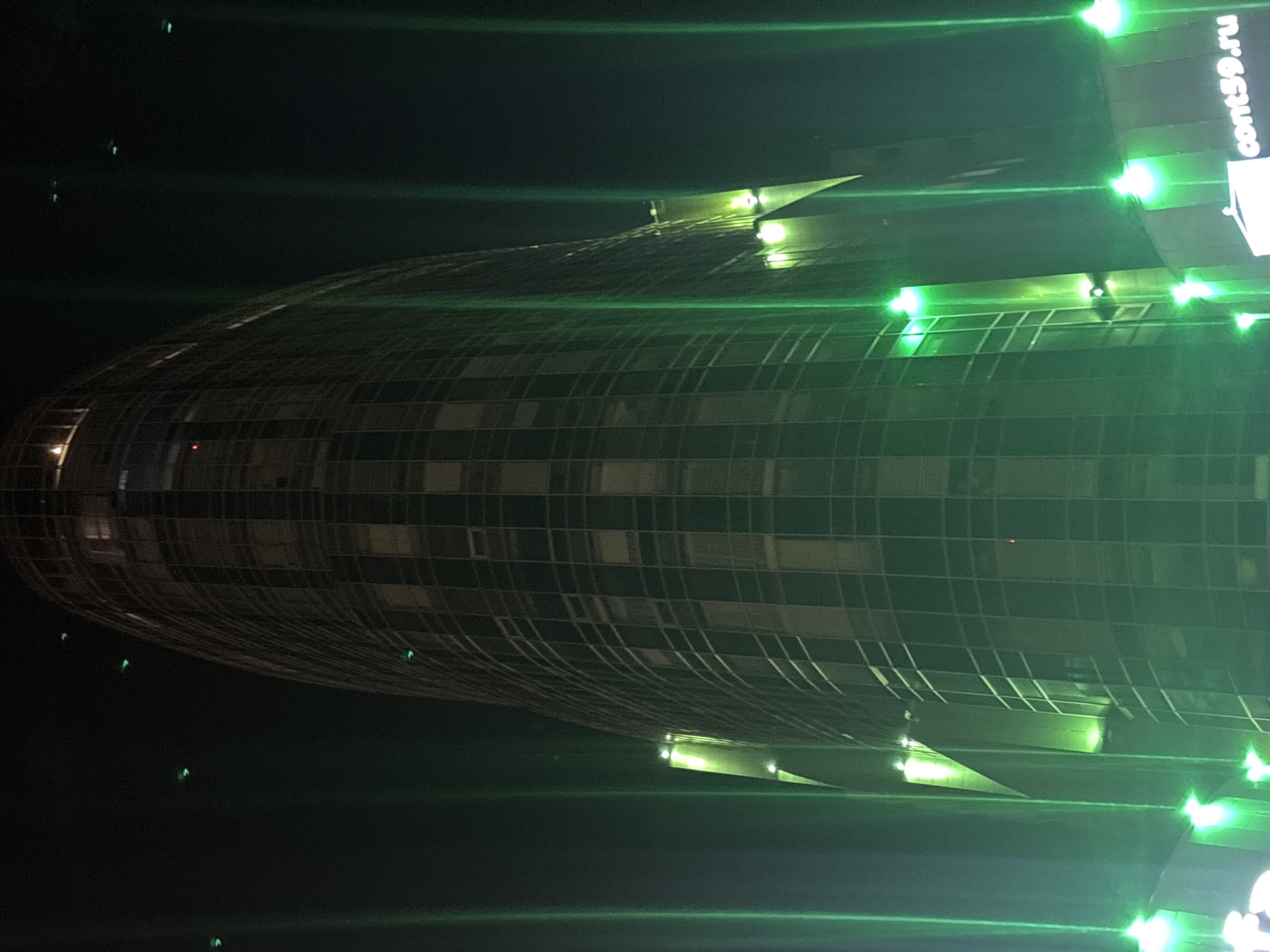 At night, a horizontally-oriented, low-angle, medium shot reveals a curved, grid-like structure. The structure is illuminated with green light and appears to be a building. The text "cont59.ru" is visible in the upper right corner. The presence of Russian domain in the text suggests the location is likely in Russia. Given the context of being located in Perm, Russia, the illuminated building is likely the Perm Regional Multifunctional Center. The surrounding environment is dark, suggesting an urban setting. The image has a grainy appearance.