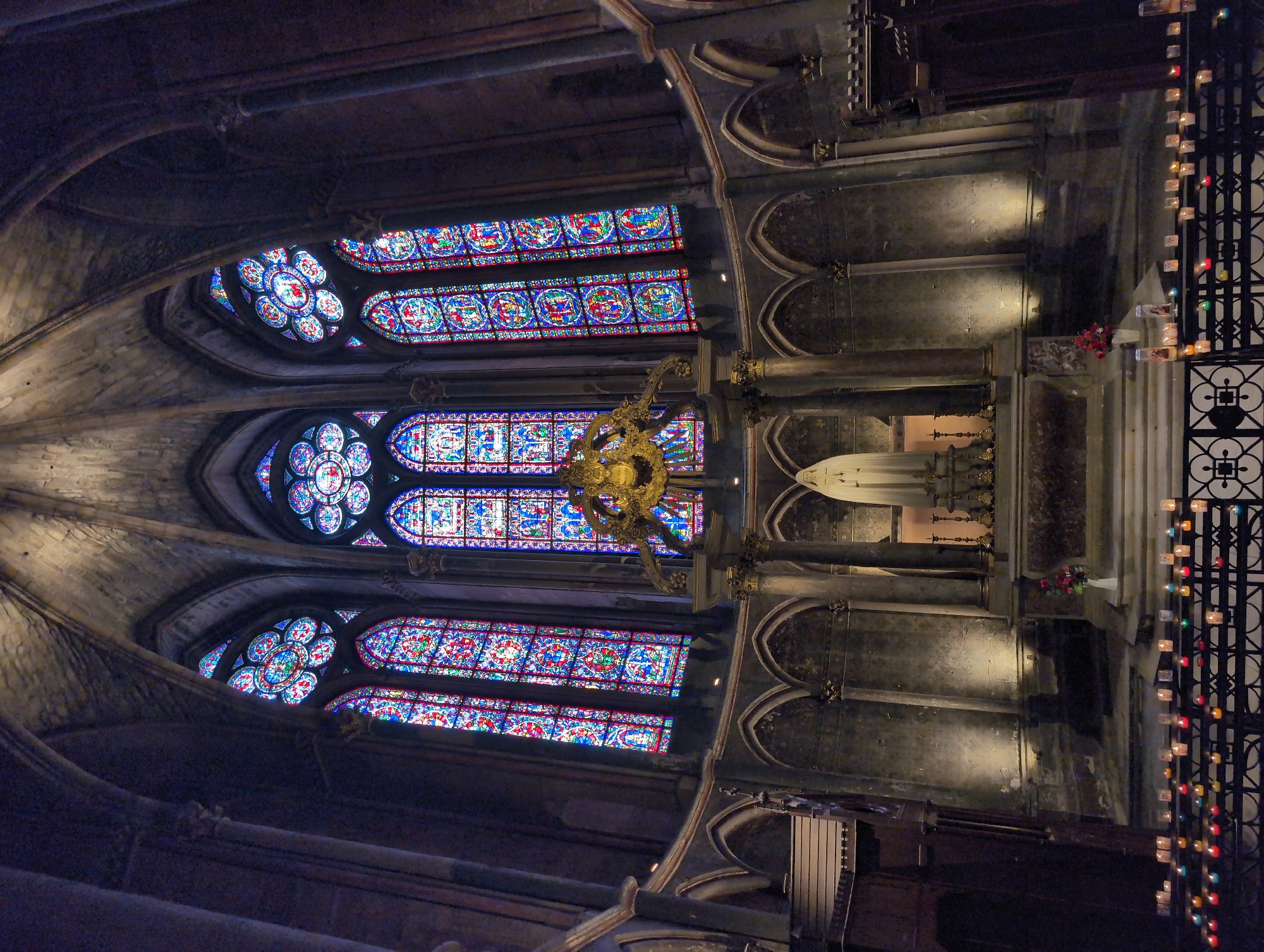 This image depicts the interior of a grand cathedral, likely a place of worship, showcasing its magnificent stained-glass windows and an altar. The cathedral's architecture features high vaulted ceilings and intricate stonework, creating a somber and awe-inspiring atmosphere. In the center, a white statue of a religious figure stands on a raised platform, adorned with flowers and lit by spotlights. In front of the altar, numerous small candles are arranged on a black metal railing, casting a warm, flickering glow. The stained-glass windows, illuminated from behind, display vibrant colors and detailed biblical scenes, adding to the ethereal beauty of the space. There are no people visible in the image, and the scene is static, suggesting a moment of quiet contemplation or prayer. The overall impression is one of reverence and historical grandeur. The provided information states the location is Urvillers, France; however, the architectural style and grandeur of the stained glass windows strongly suggest a much larger and more famous cathedral, such as Notre Dame de Paris, which is a more fitting depiction of such a magnificent structure.
