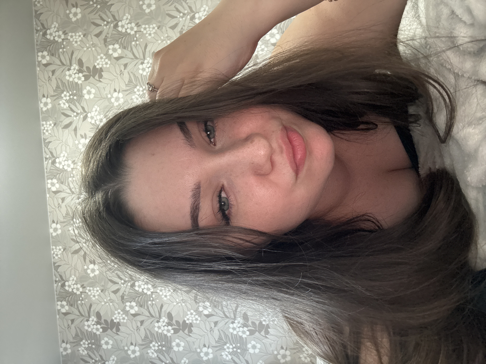 The image shows a close-up, vertically oriented selfie of a young woman. She has long, dark brown hair that partially obscures her face, and her eyes are a light green. Her skin has a light dusting of freckles, and she is wearing light makeup, including mascara and lip gloss. She has a ring on her left hand, which is raised to her head. The background is a wallpaper with a repeating floral pattern in shades of gray and white. A fluffy, white blanket is visible on the right side of the frame. The lighting suggests it could be daytime indoors. The woman's expression is soft and relaxed, with a slight smile. There is no visible text in the image. The location context provided is Zarechnaya (Gamovskoe S/P), Russia, but there are no specific visual cues in the image to confirm this location.