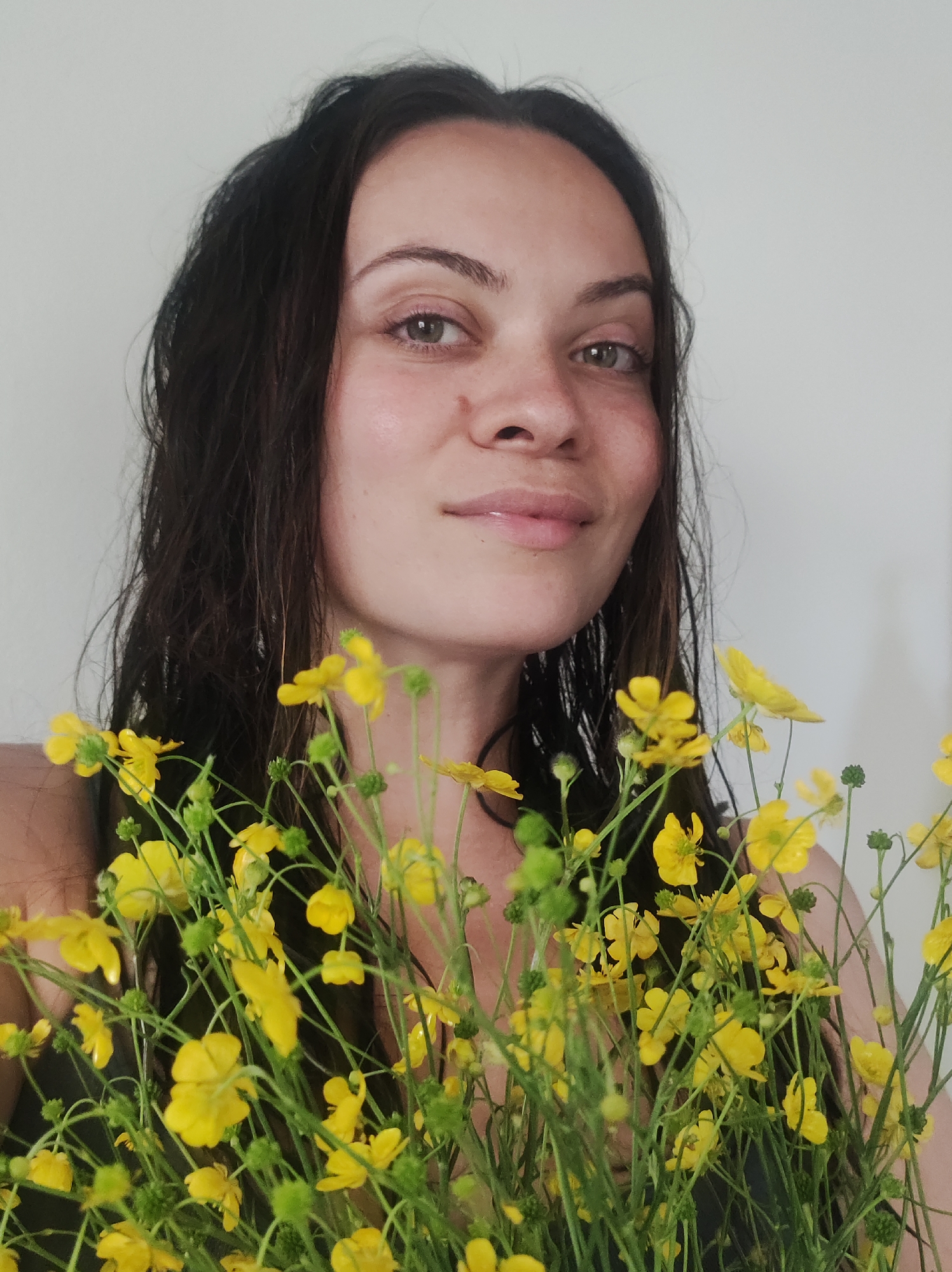 A head and upper-torso portrait captures an adult female holding a dense cluster of yellow buttercup flowers in an interior setting, city of Unknown, Unknown. The female subject, positioned centrally, has long, dark, wet-appearing wavy hair cascading over her shoulders. Her face is directed towards the viewer, displaying a slight, closed-mouth smile. Her eyes are light-colored, possibly hazel, with visible pupils. A small reddish mark or scar is present on her right cheek, below the outer corner of her eye. She wears dark, possibly black, attire, with a portion of a shoulder strap visible on her left side. The vibrant yellow buttercups, characterized by numerous small blooms and green stems, occupy the lower half of the frame, partially obscuring her lower face and chest. The background is a plain, light-colored, untextured wall. The lighting is soft and even, highlighting the subject's features and the flowers' details.