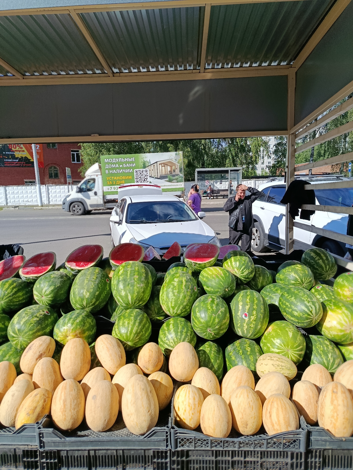 This media file shows an outdoor fruit stand on a sunny day, likely in Orekhovo-Zuyevo, Russia. The foreground is dominated by a large display of watermelons and melons, neatly arranged in black crates. Some watermelons are cut in half, revealing their bright red flesh. Behind the fruit stand, a street scene unfolds. Several white vehicles, including a van and a car, are parked or driving on the road.
A man is prominently featured in the mid-ground, standing by a white car and talking on his phone, appearing somewhat flustered or stressed as he holds his hand to his forehead. In the background, there are more cars, a bus stop shelter, and buildings, suggesting an urban or suburban environment. Trees with green foliage line the street, indicating it's likely summertime.
Visible text on a banner behind the fruit stand advertises "MODULAR HOUSES AND BATHS AVAILABLE. WE WILL INSTALL IN 3 DAYS. +7 93" and a website address "goodwoodhouse.ru". Another banner in the background displays a safety warning about fire hazards. The overall impression is a lively, everyday scene of commerce and street life on a warm day.